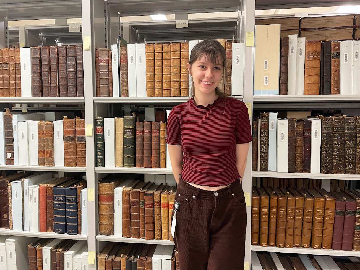 Kate Oruc, student assistant at William Ready Division of Archives and Research Collections, stands smiling in front of shelves of rare books.  