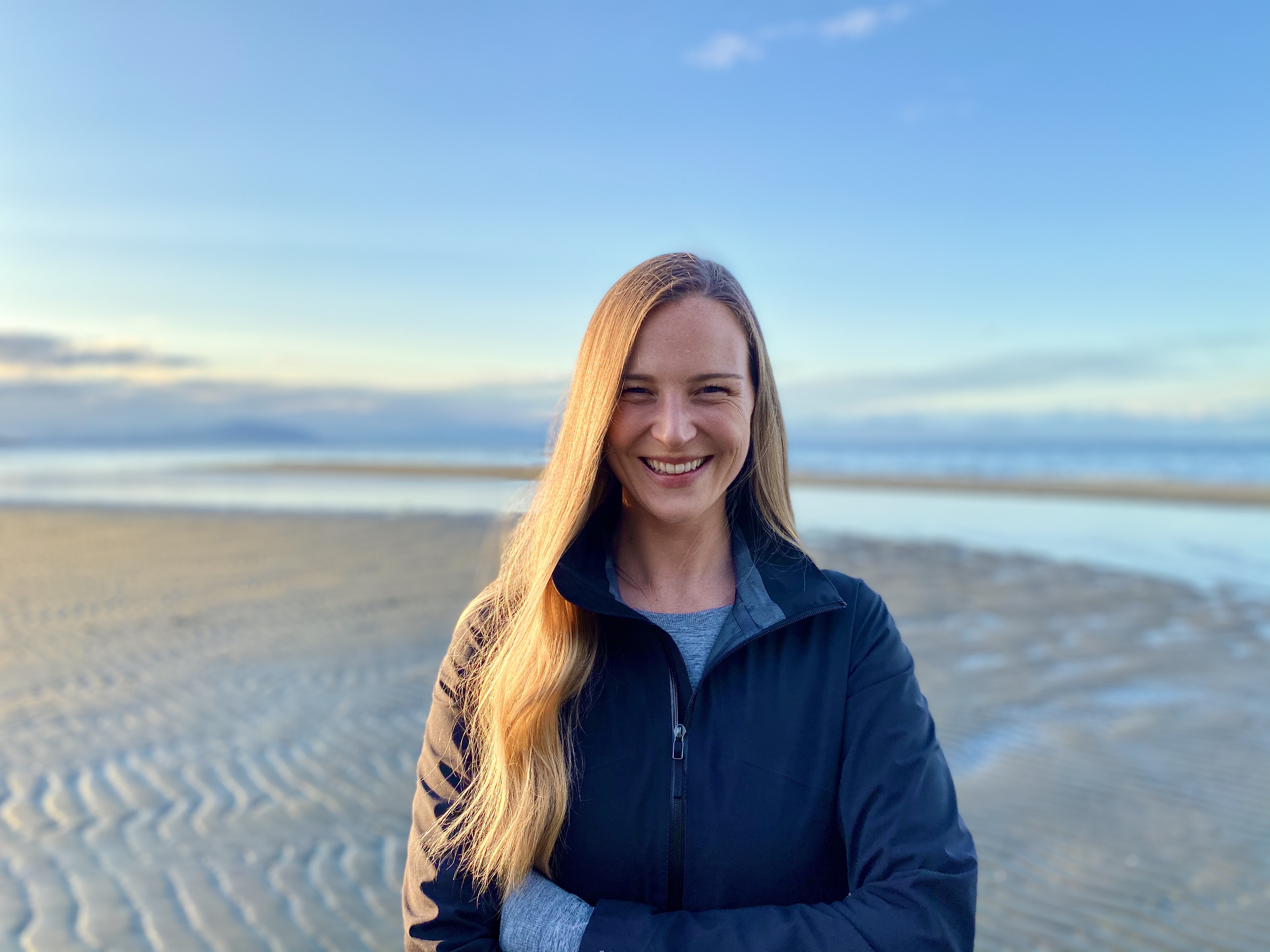 Alix Shield with long blonde hair wearing a navy jacket and grey sweater stands smiling outdoors on a sandy beach in front of a large body of water. 