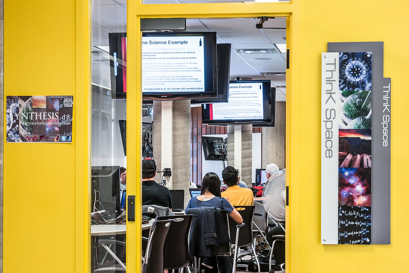 The entrance to the ThInk Space on the third floor of Thode Library. The door is a bright yellow and a sign on the right says the name of the space. Beyond the open door are students at tables with large monitors hanging from the ceiling displaying information for them.