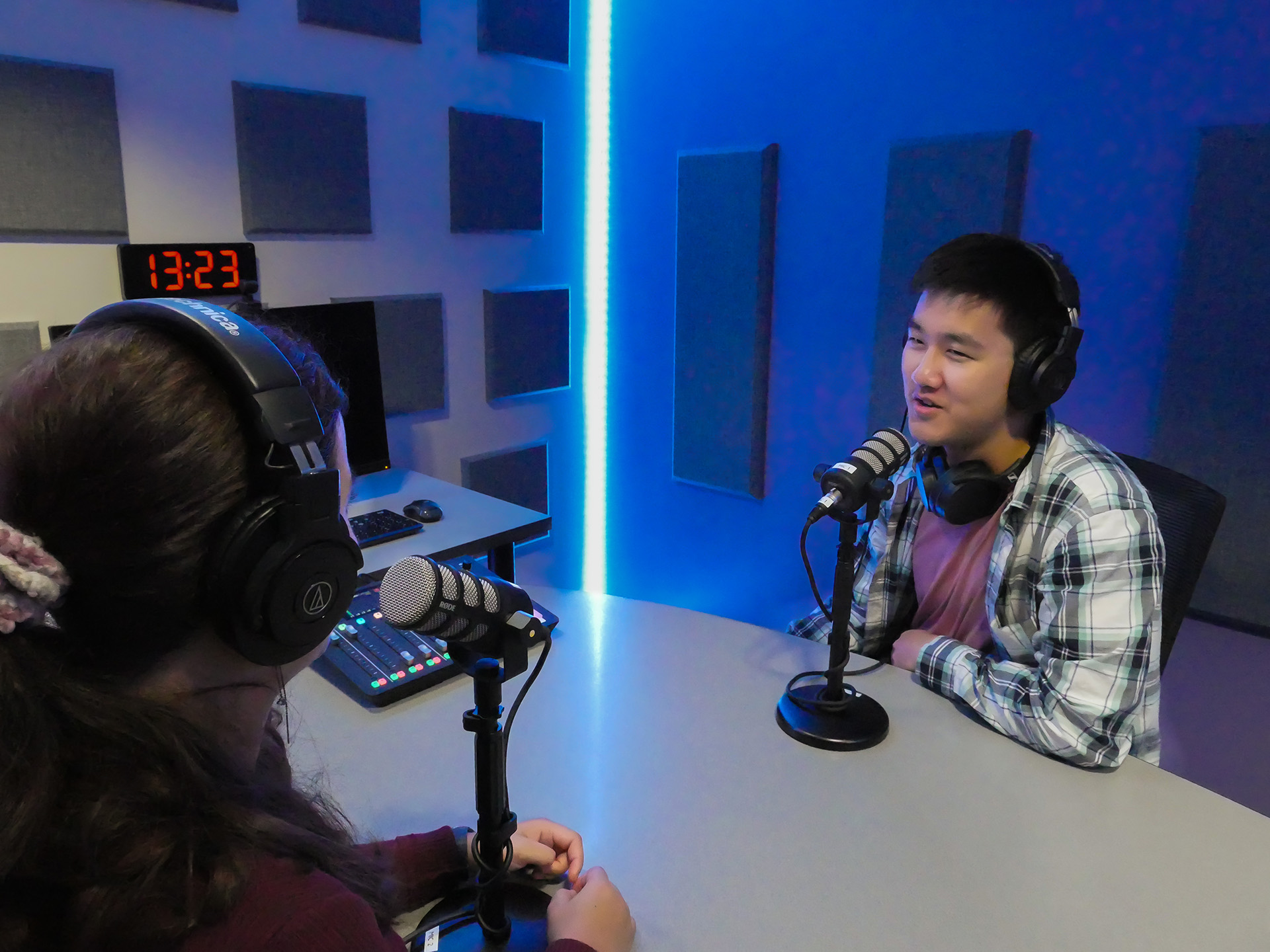 Two students sit at a table in the Podcast Studio in Lyons, wearing headphones and speaking into podcasting microphones. Soundproofing panels line the walls and LED lights are lined up along the corner of the room from floor to ceiling.