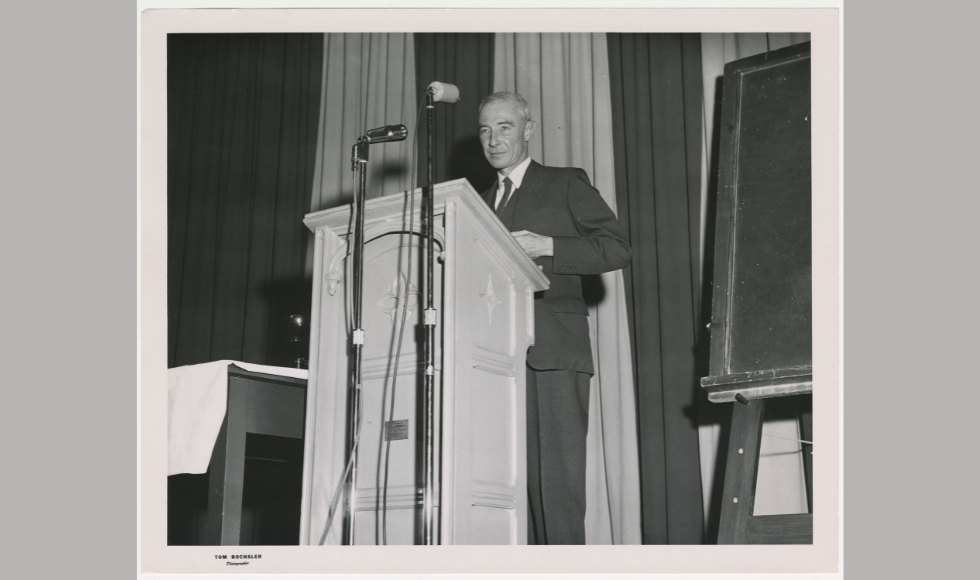 A black and white archival photo shows a person with white hair and wearing a suit at a podium at McMaster. 