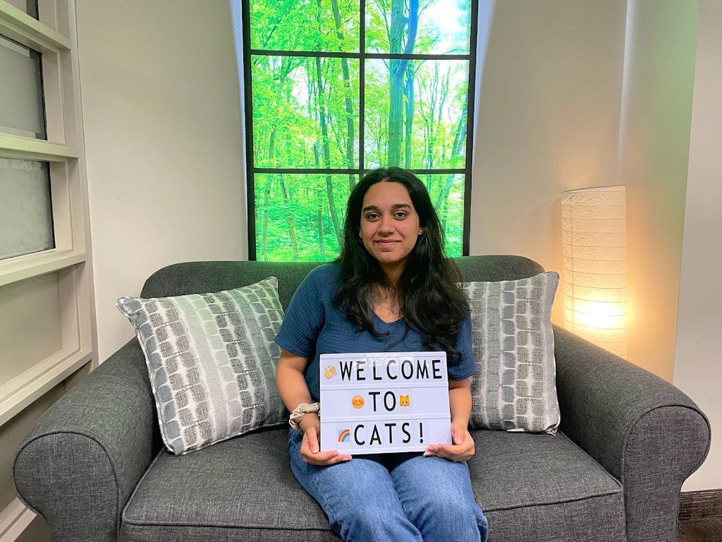 Manarpan Kaur, transcription assistant at Library Accessibility Services, can be seen sitting on a couch inside the Campus Accessible Tech Space and holding a sign that reads Welcome to CATS!