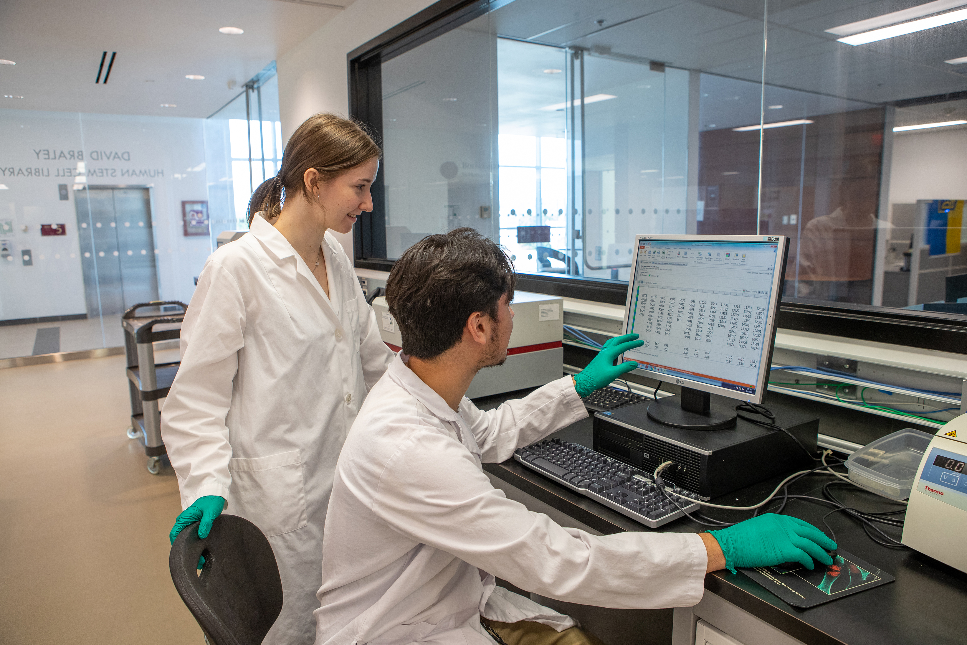 Two students wearing white lab coats sit at a computer inside a laboratory analyzing data on a computer screen. 