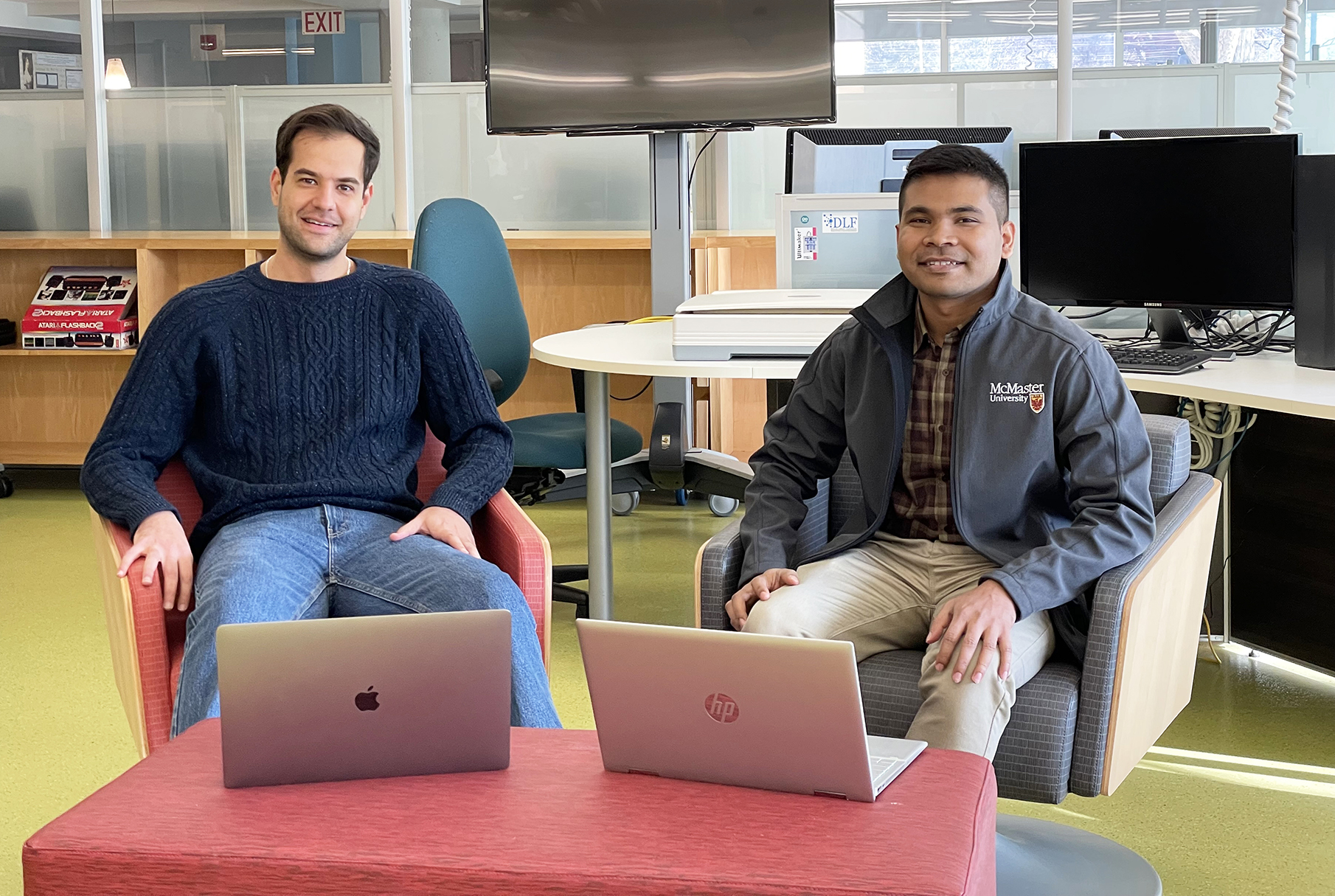 Seyed with black hair wearing a blue sweater and Humayun with black hair and a grey sweater sit together inside Sherman Centre with their laptops on the coffee table in front of them. 