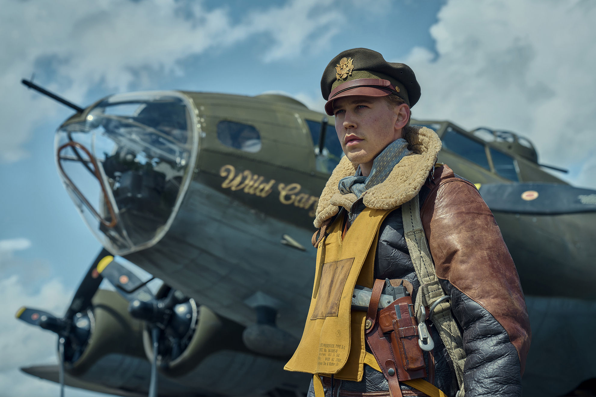 Austin Butler wearing Second World War bomber jacket with a yellow parachute vest stands besides a vintage airplane on a movie set. 