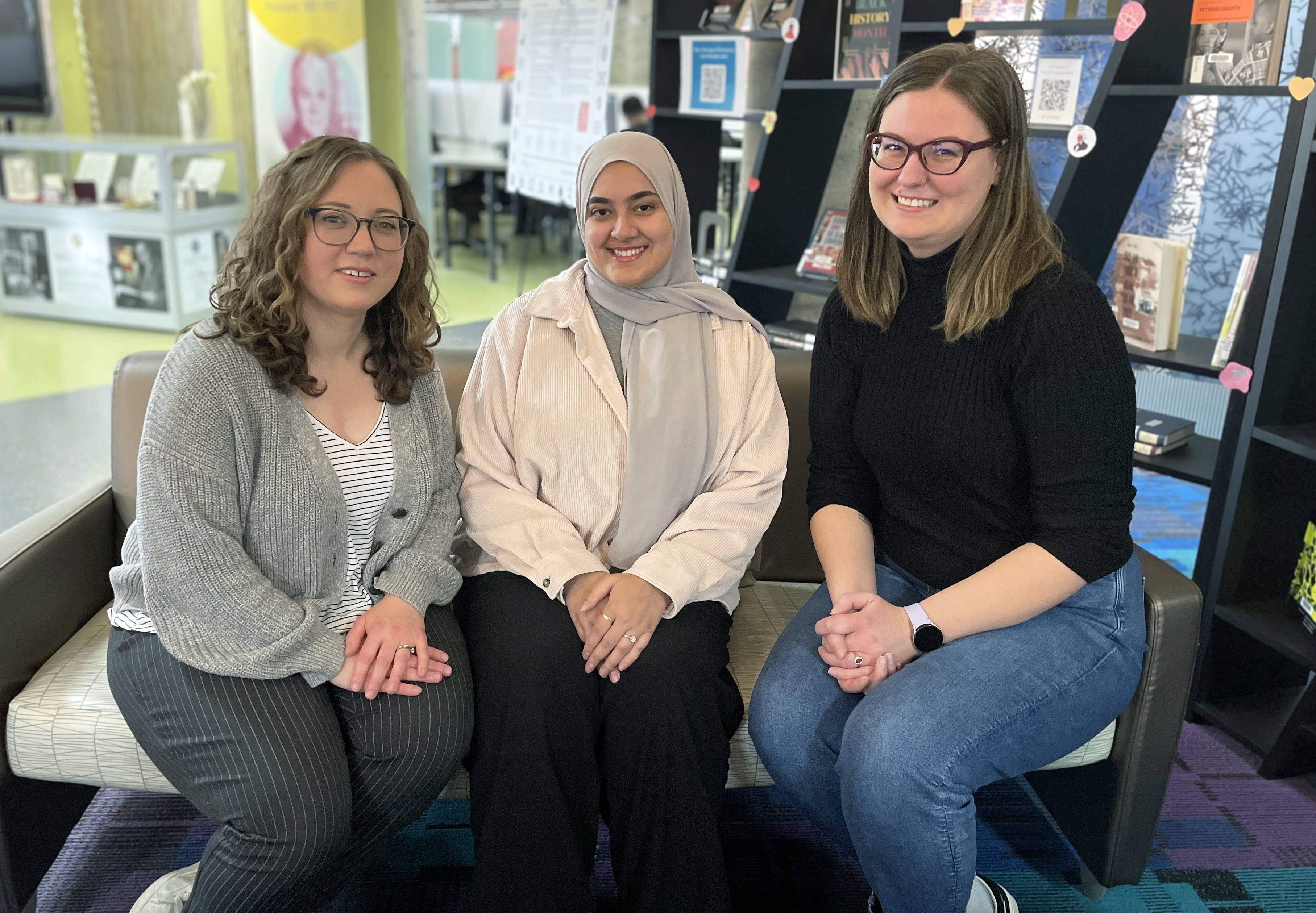 Three staff members from McMaster University Libraries’ Teaching and Learning Mini Conference planning committee sit on a couch indoors smiling near a book display. 