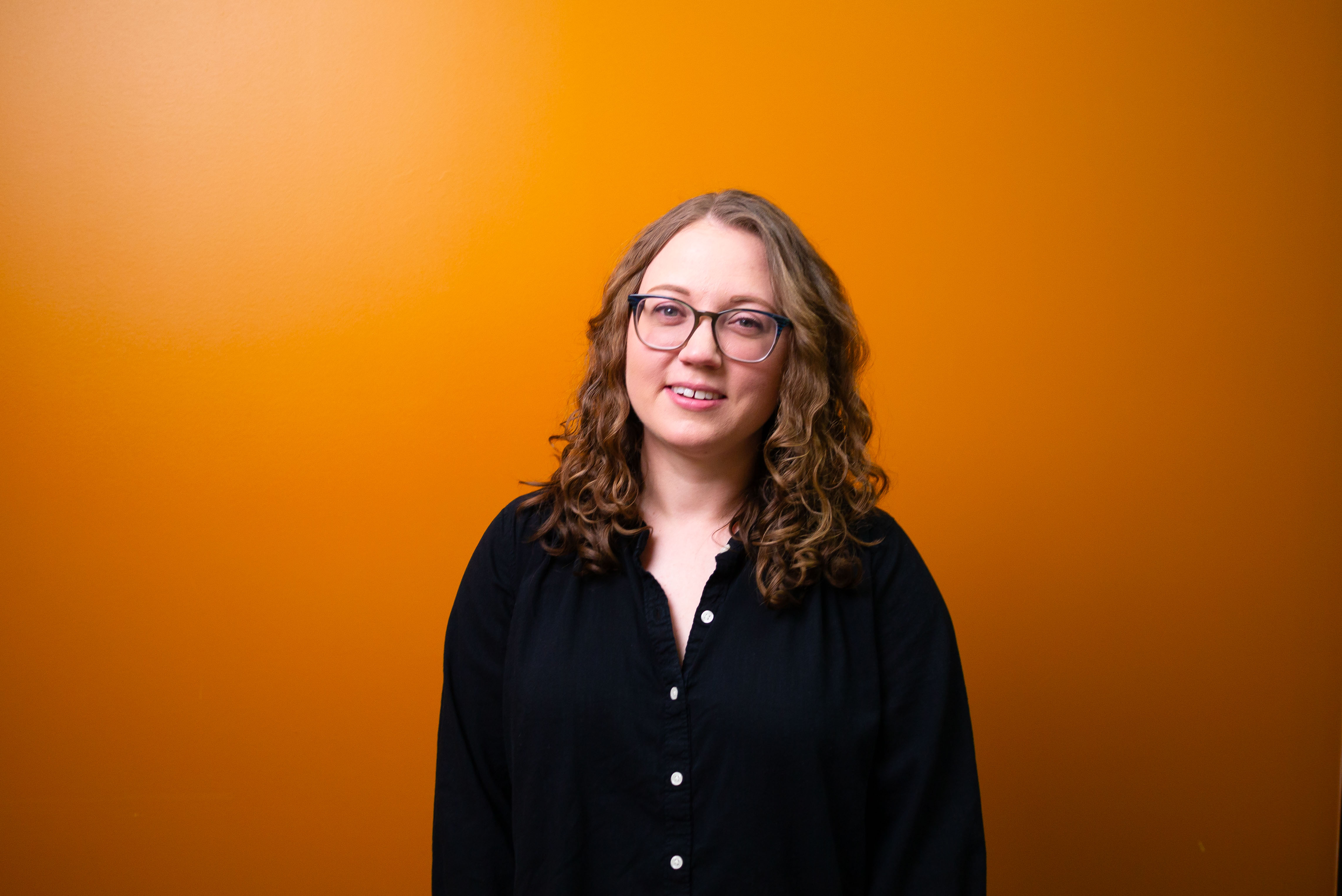 Katie Harding with long brown curly hair wearing glasses and a black button-up blouse stands smiling indoors in front of an orange wall. 
