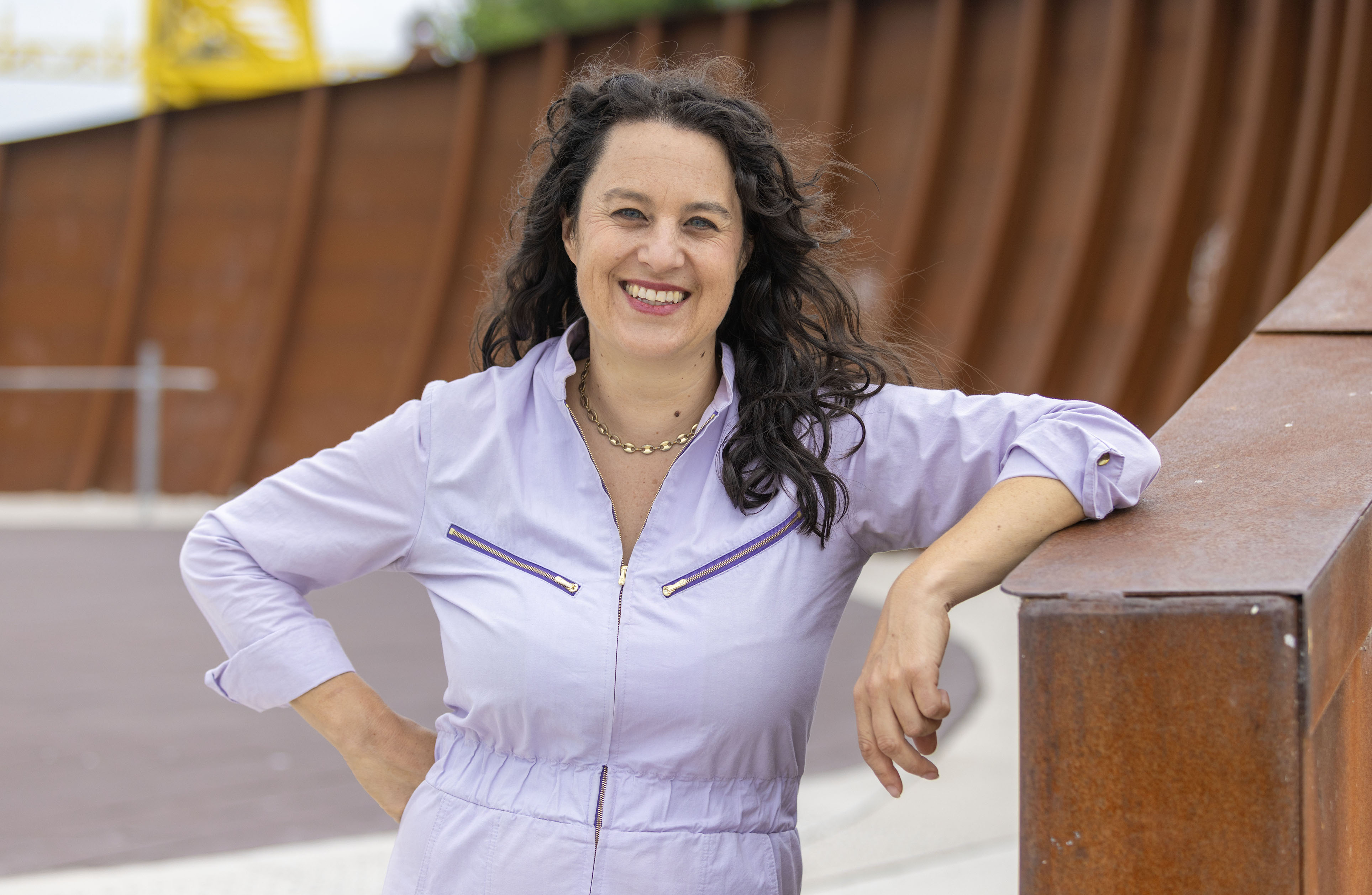 Anna with long black hair wearing a light purple blouse stands smiling outdoors.
