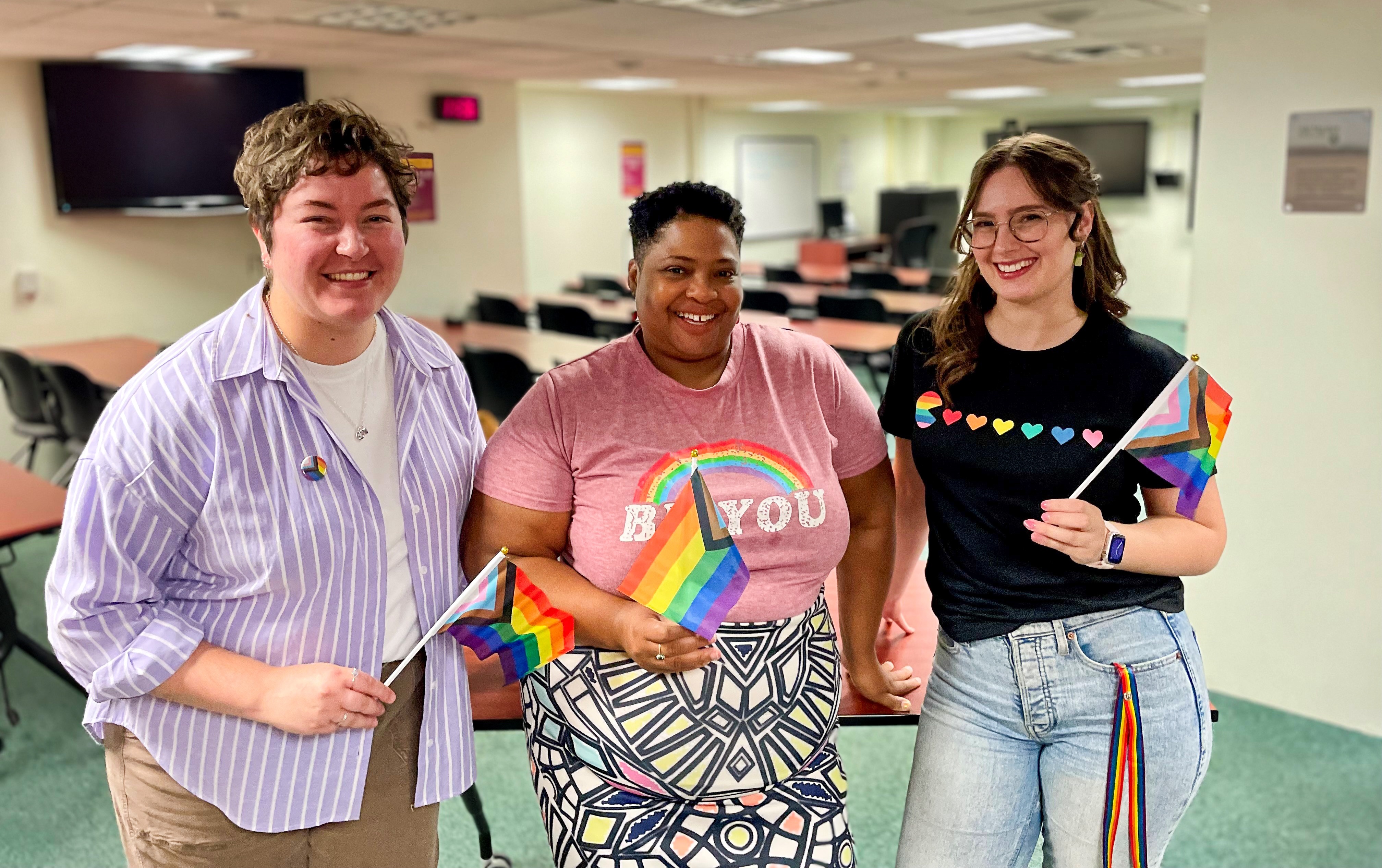 Three library staff members standing and smiling, holding pride flags.