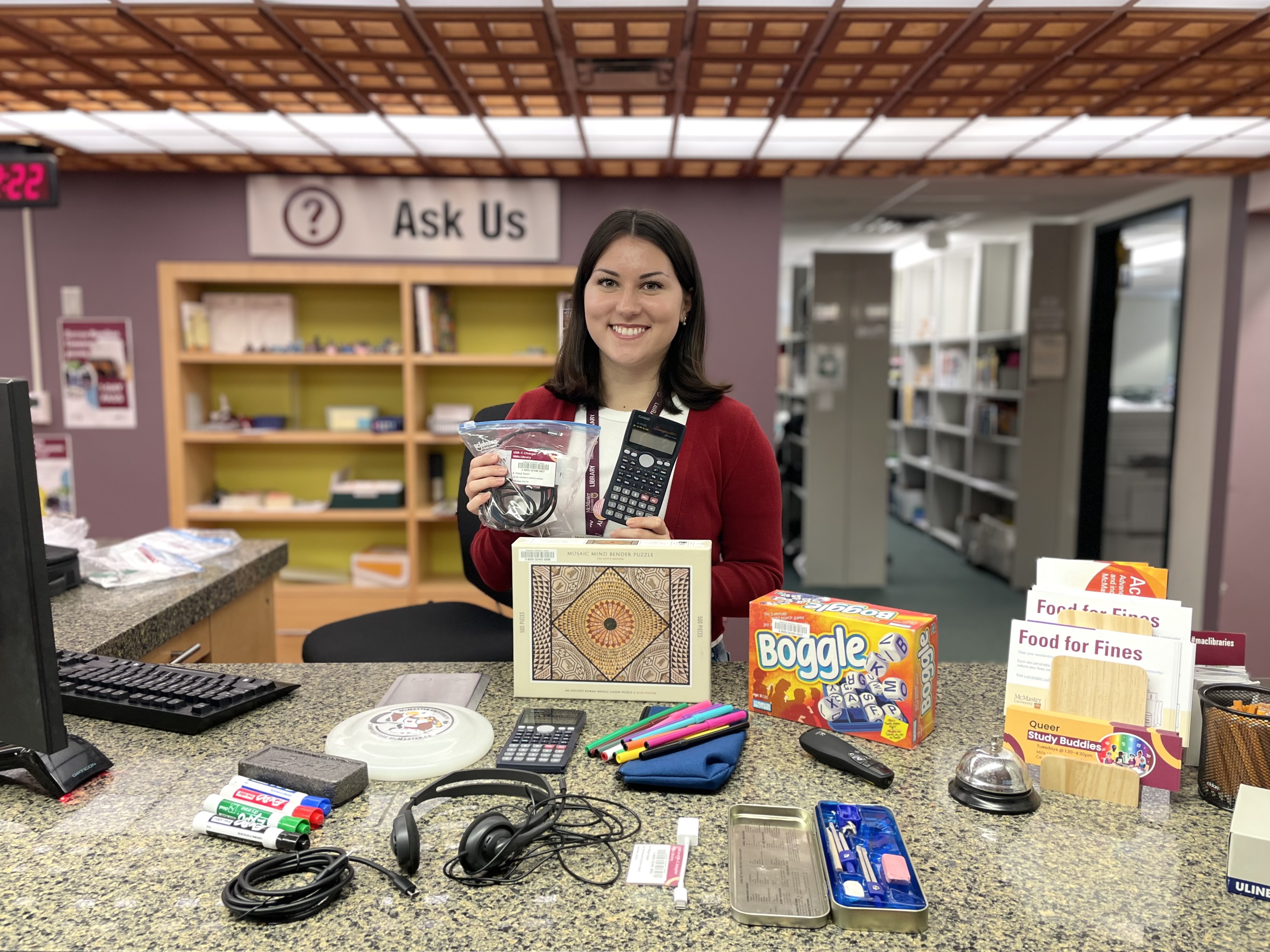 Grace with shoulder-length black hair wearing a red cardigan stands smiling behind the services desk in Mills library. Grace holds a calculator in one hand and a bag with charging cables in the other hand, with a variety of items laid out on the desk in front of her.