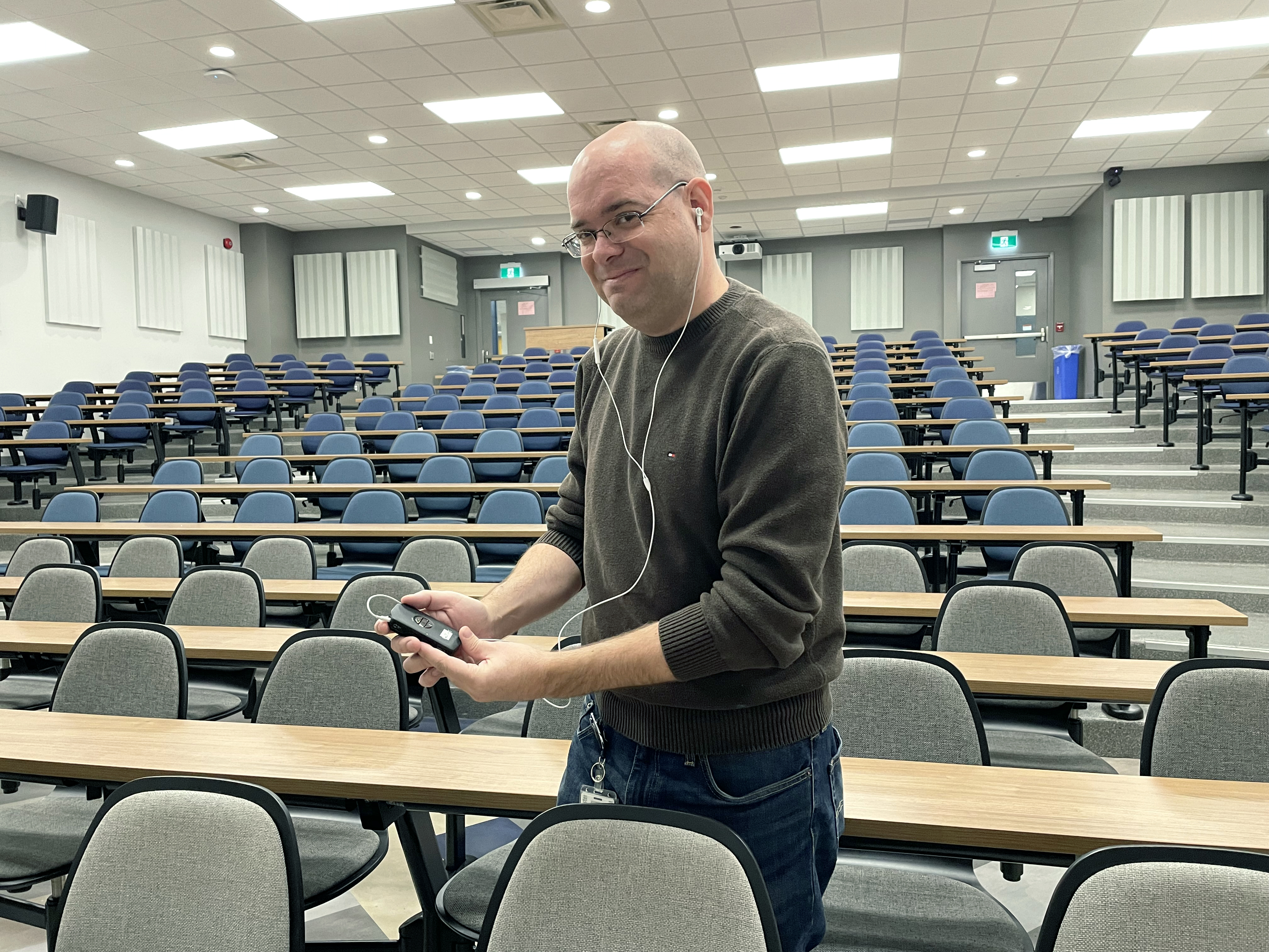 Joey with a shaved head wearing glasses and a brown sweater stands in a lecture hall with rows of blue and grey chairs. He is wearing earbuds which are connected to the assistive listening device he is holding.