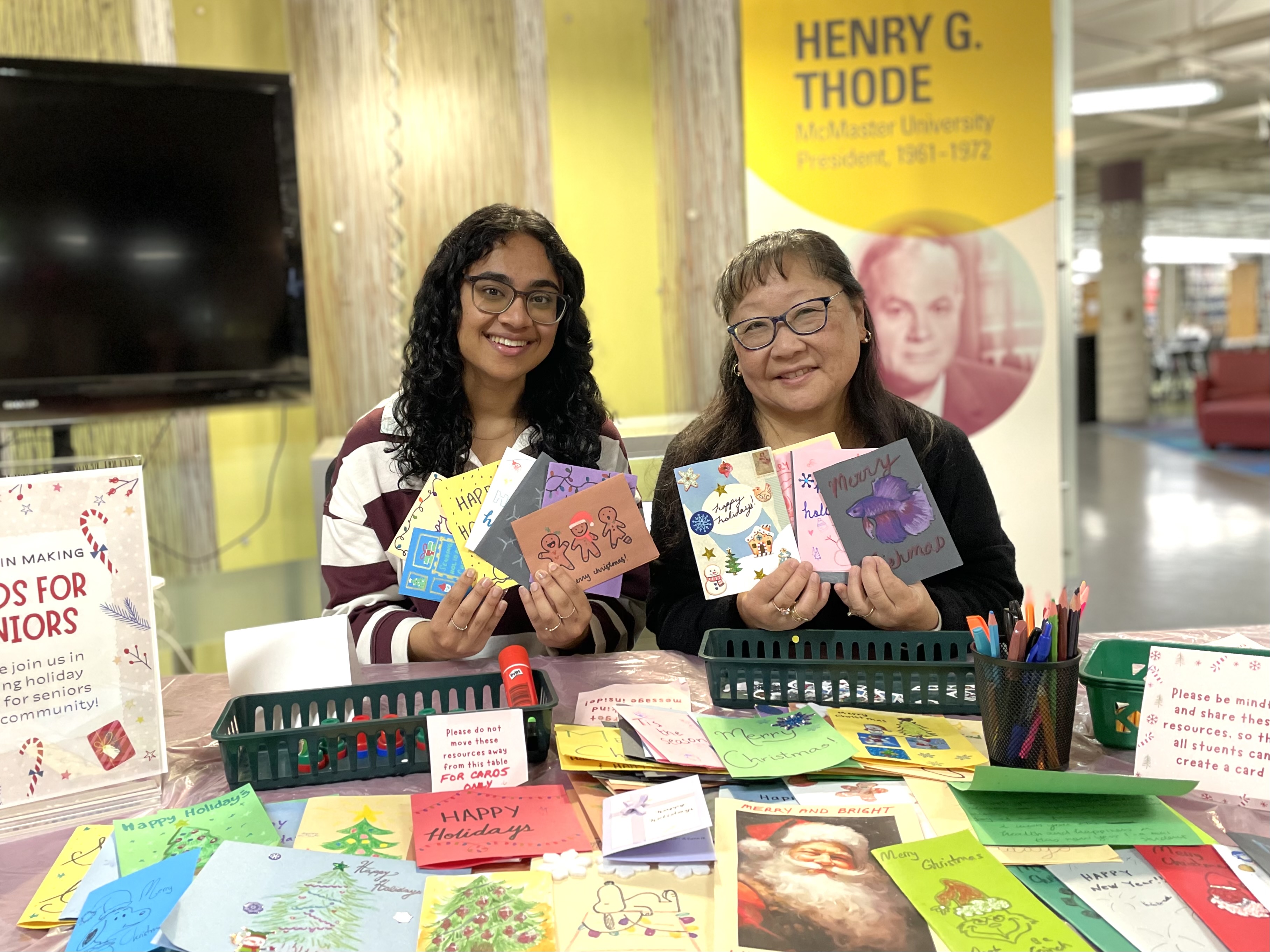 Poornima with long black hair and glasses and Ann with long black hair and glasses sit at the holiday card for seniors table in H.G. Thode Library holding cards made by students.