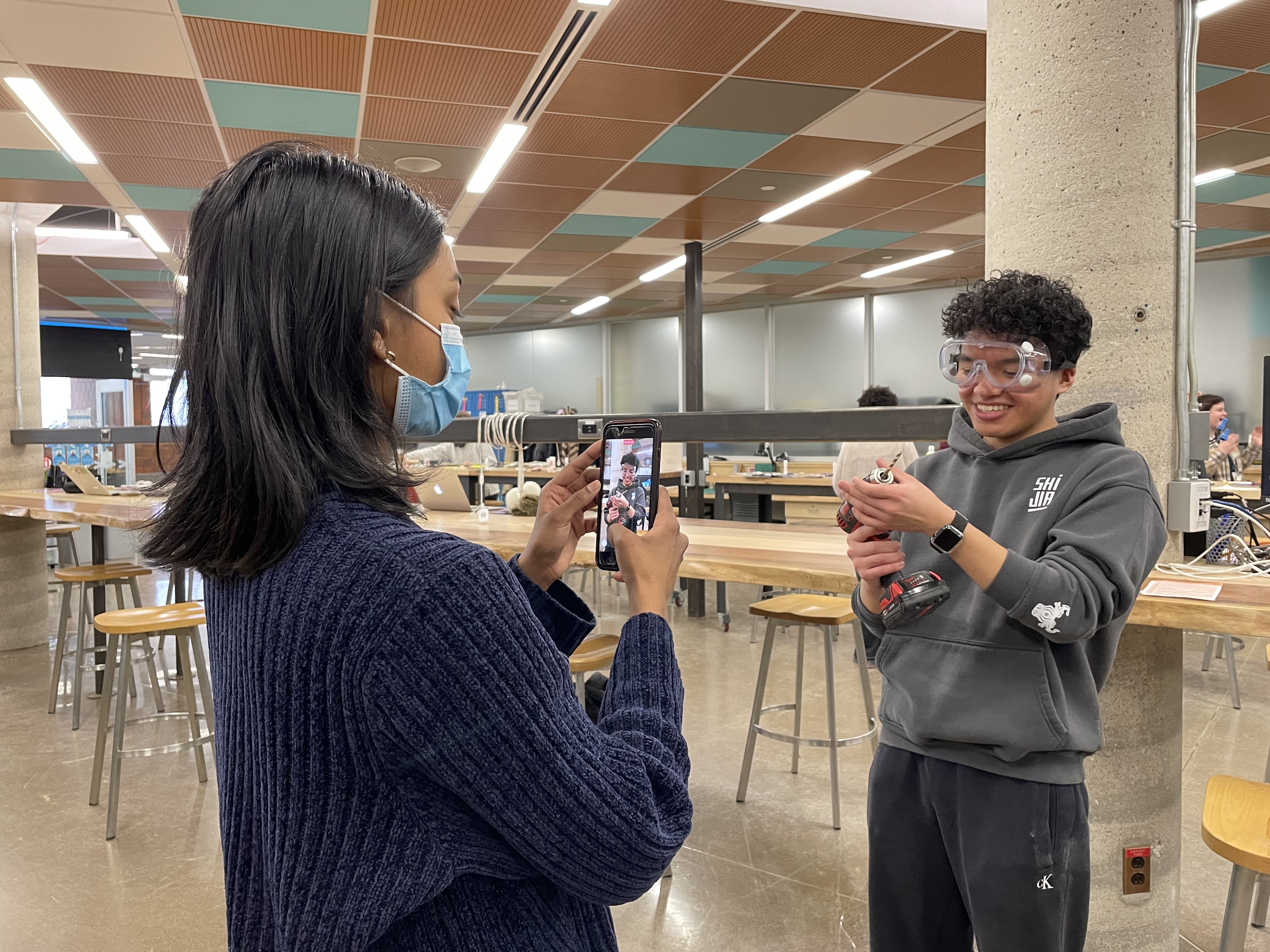Meenaa with long black hair wearing a blue sweater holds a cell phone to film Dominick. Dominick with short black hair and a grey sweater stands in front of Meenaa holding a hand drill.