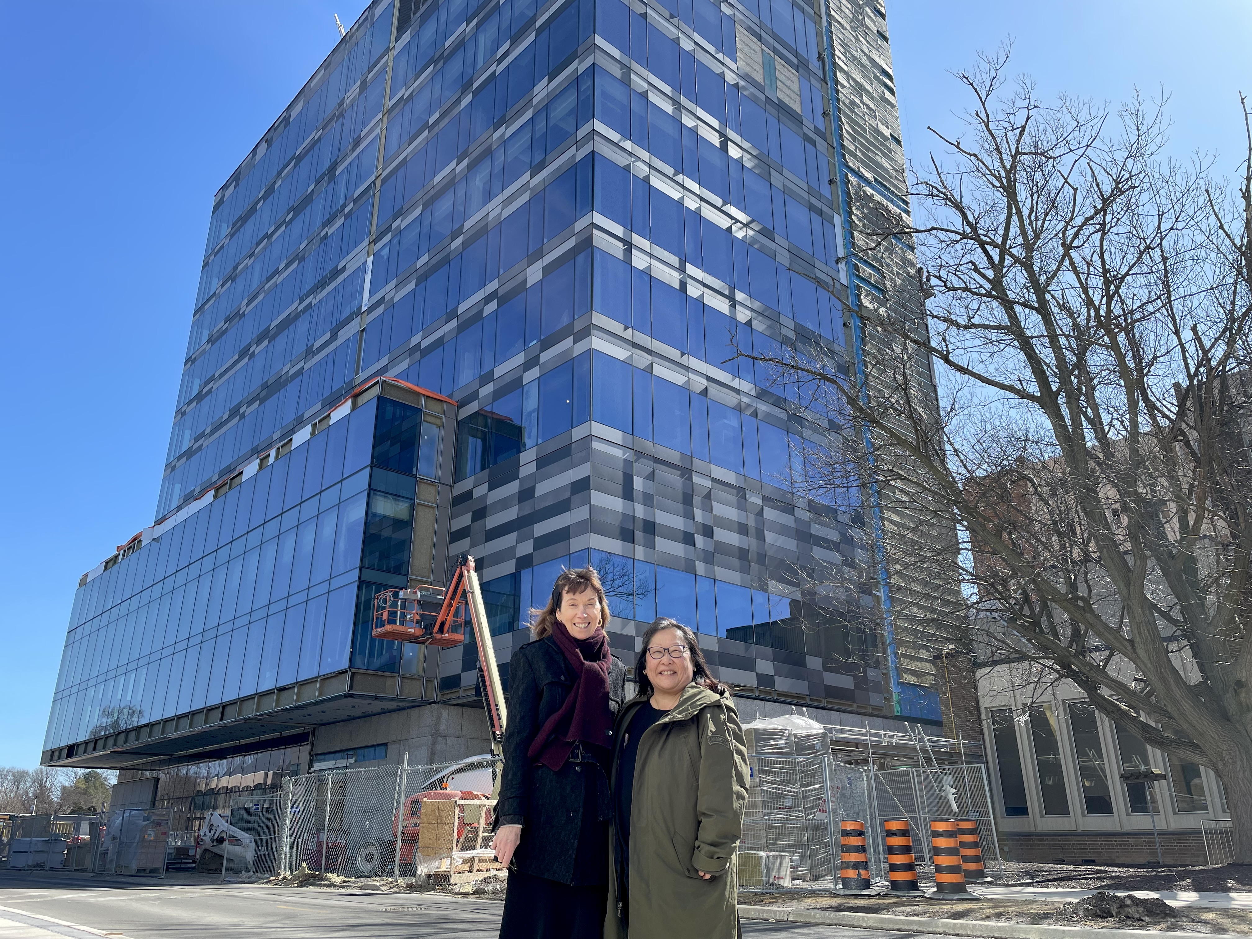 Vivian with long brown hair wearing a black coat and a scarf and Ann with long black hair and glasses wearing a green coat stand together smiling outside of the McLean Centre for Collaborative Discovery on a sunny day.
