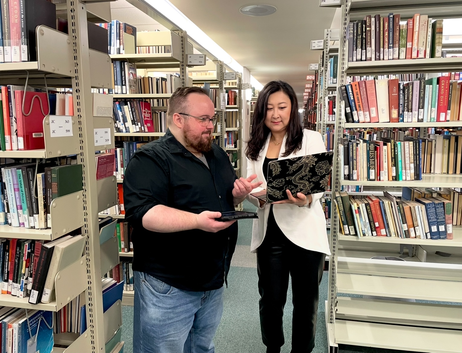 Cameron Wheaton with short brown hair holding a tablet stands next to Wei Zhang with long black hair holding a laptop in Mills Memorial Library.