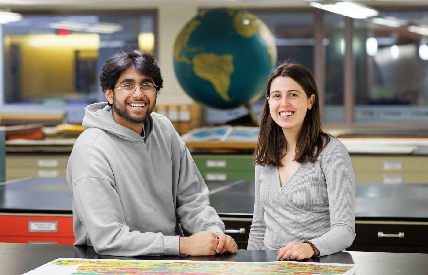 Dharav with short black hair wearing glasses and a grey hooded sweather and Esther, with shoulder-length brown hair wearing long-sleeve grey sweater both stand smiling in front of a map inside the Lloyd Reeds Map Collection.