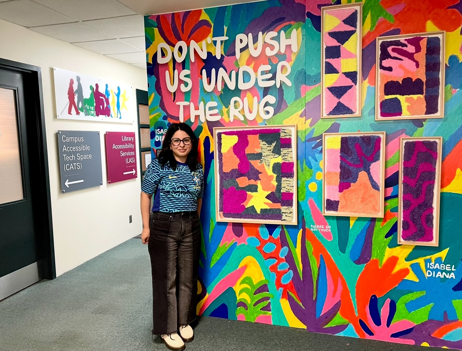 Isabel Diana with long black hair and glasses stands in front of her artwork in Mills Memorial Library.