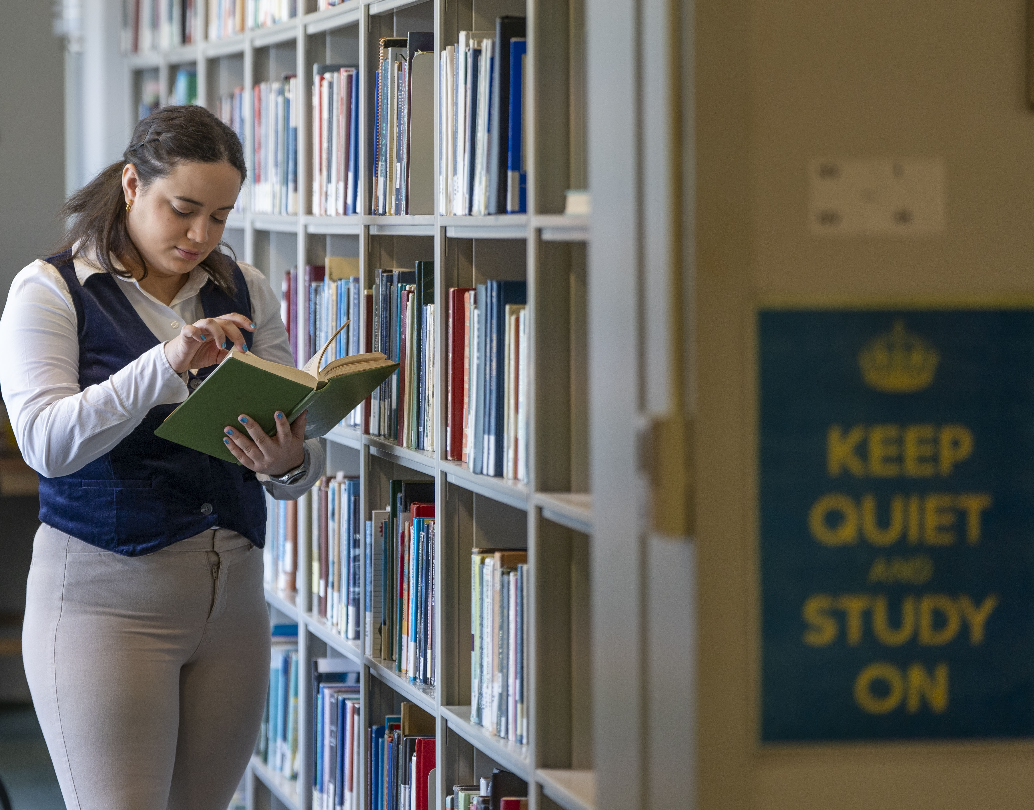 Student wearing a white dress shirt, navy vest and khaki pants with shoulder-length brown hair reads a book by a book stack in Mills Memorial Library.