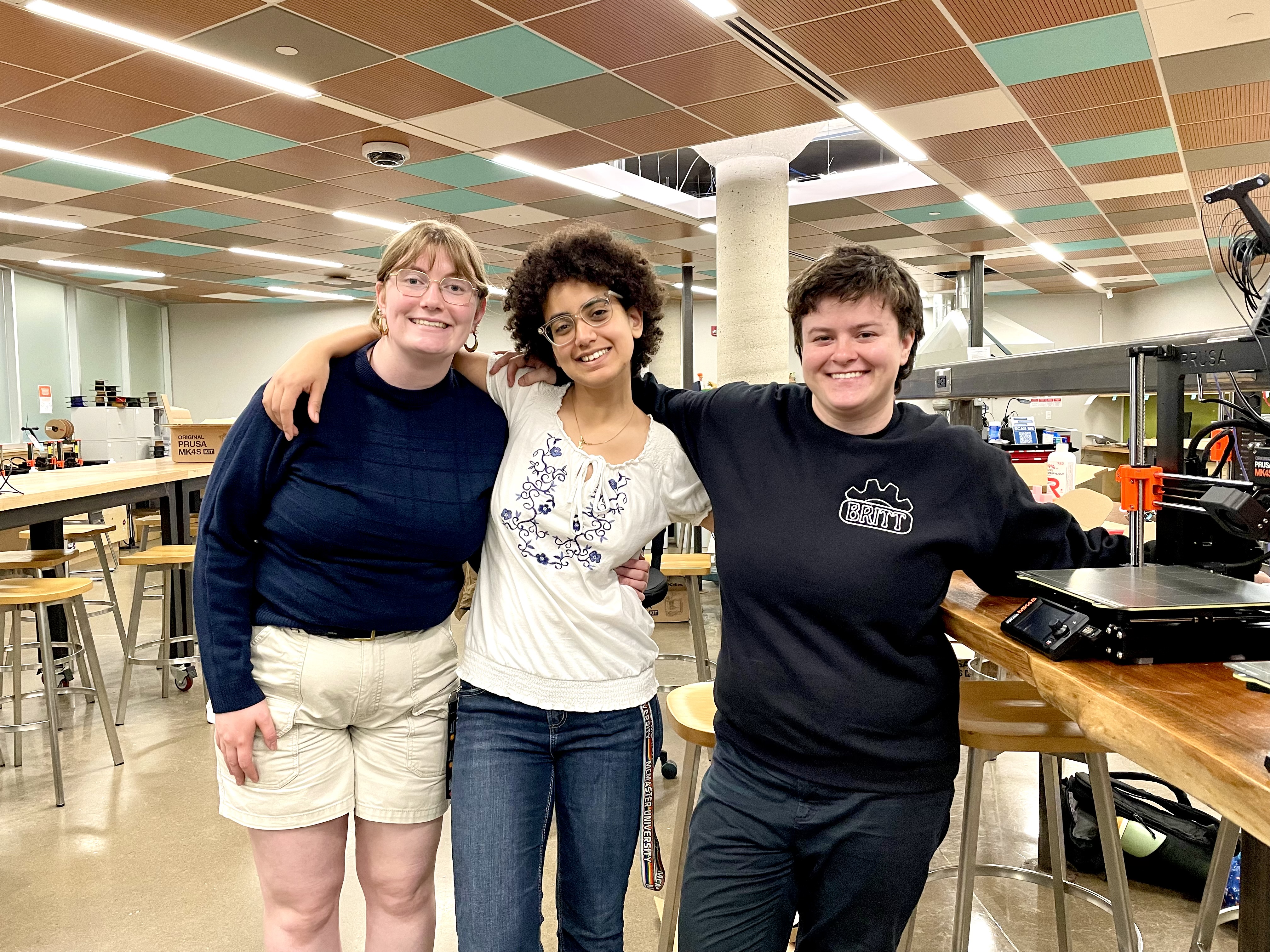 Kiana with blonde hair, glasses, and a navy sweater, Leana with brown hair, glasses and a white shirt, and Brittany with brown hair and a navy sweater stand together smiling in Thode Makerspace.