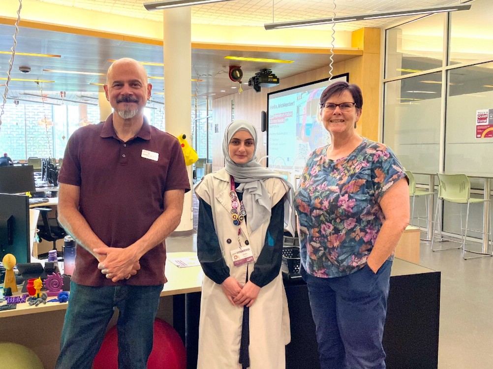 Alex with a shaved head and a maroon shirt, Zain with a blue headscarf and a white shirt, and Rhonda with short purple hair, glasses and a colourful shirt stand together smiling in Lyons New Media Centre.