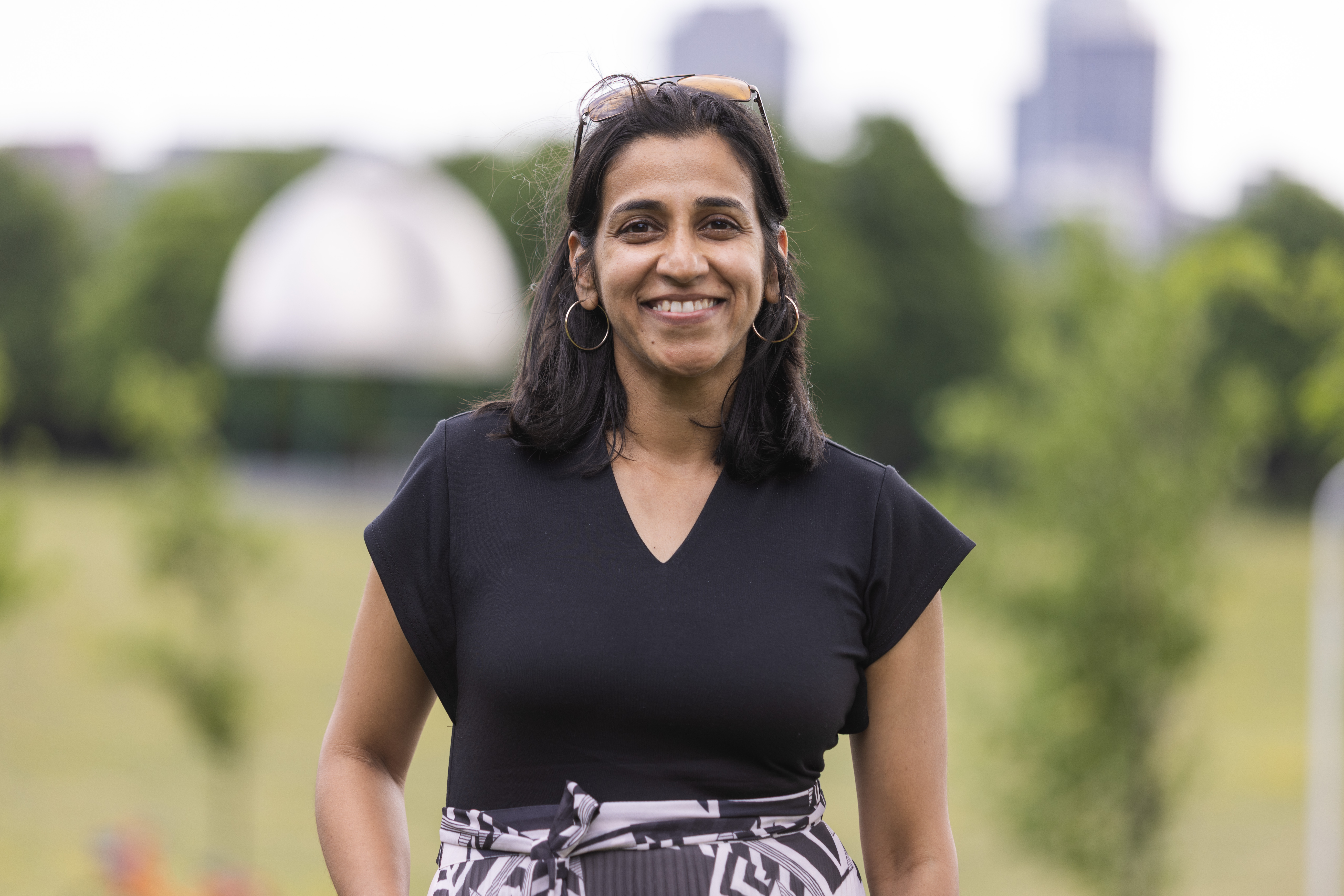 Saumiya with shoulder-length black hair wearing a black T-shirt and black and white skirt stands smiling outdoors in a park on a cloudy summer day.