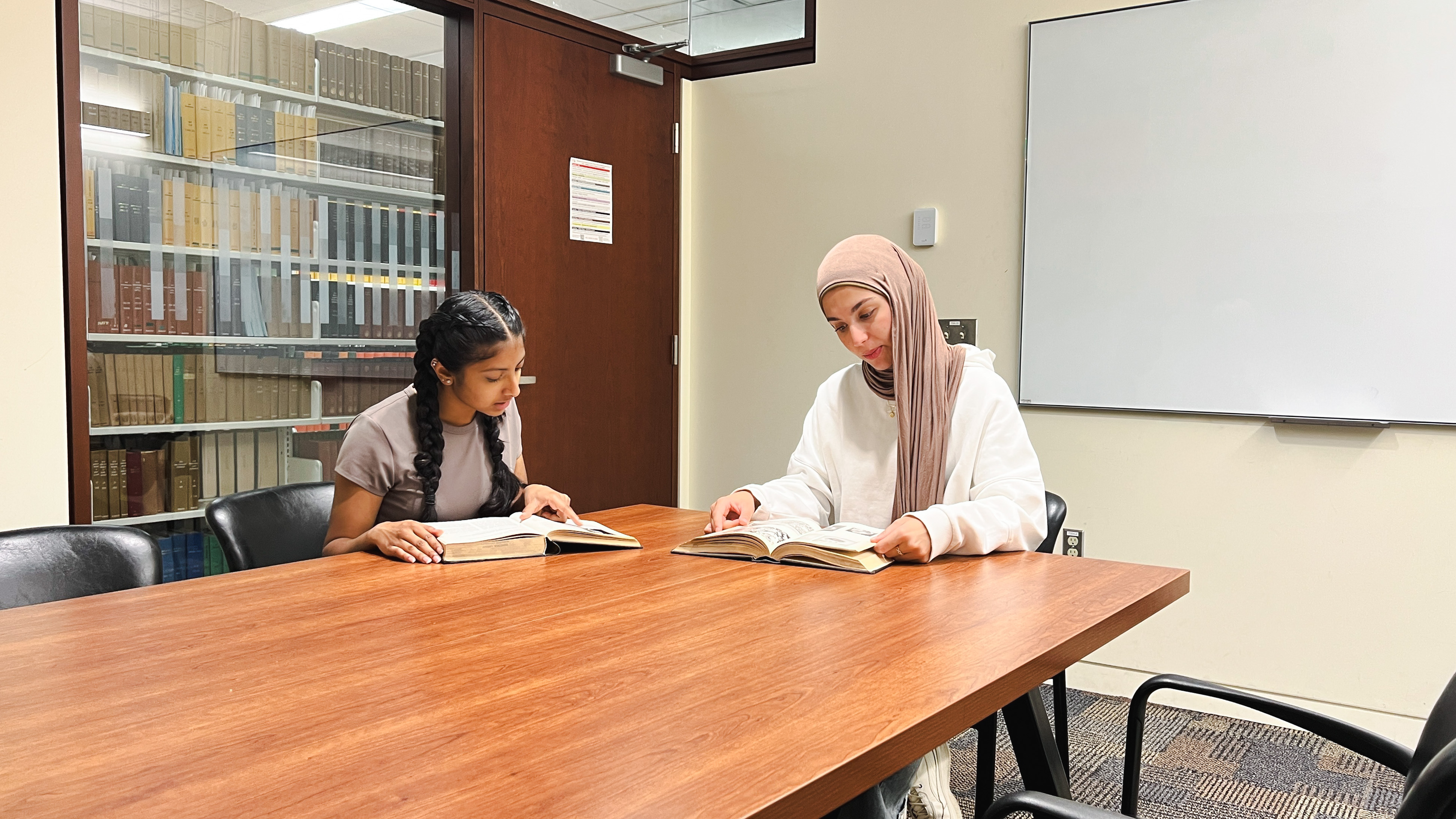 Two students sit looking at books at a table inside a group study room. 