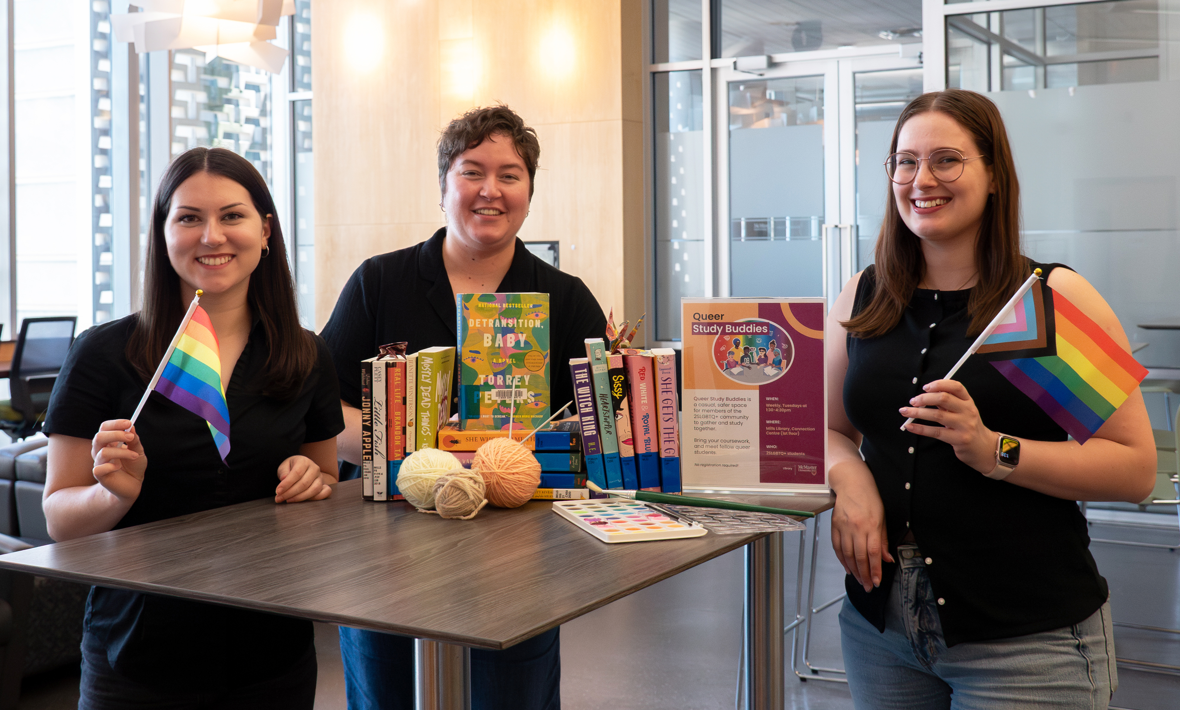 Three library employees stand smiling holding rainbow flags at a table featuring books and art supplies inside Lyons New Media Centre at Mills library. 