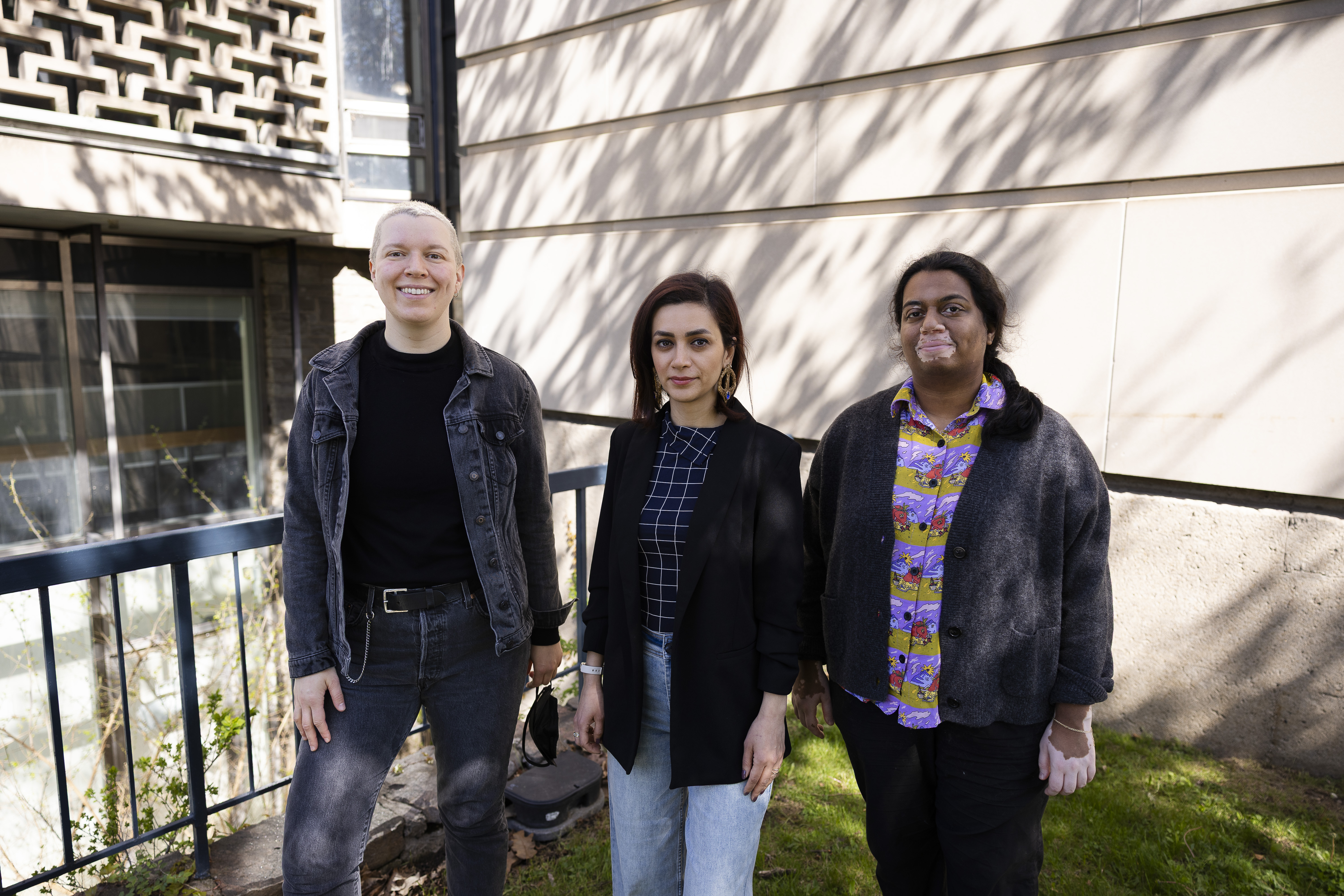 Danica with short blonde hair, Niloofar with medium-length brown hair, and Subhanya with long black hair stand smiling outside of a library on a sunny spring day.