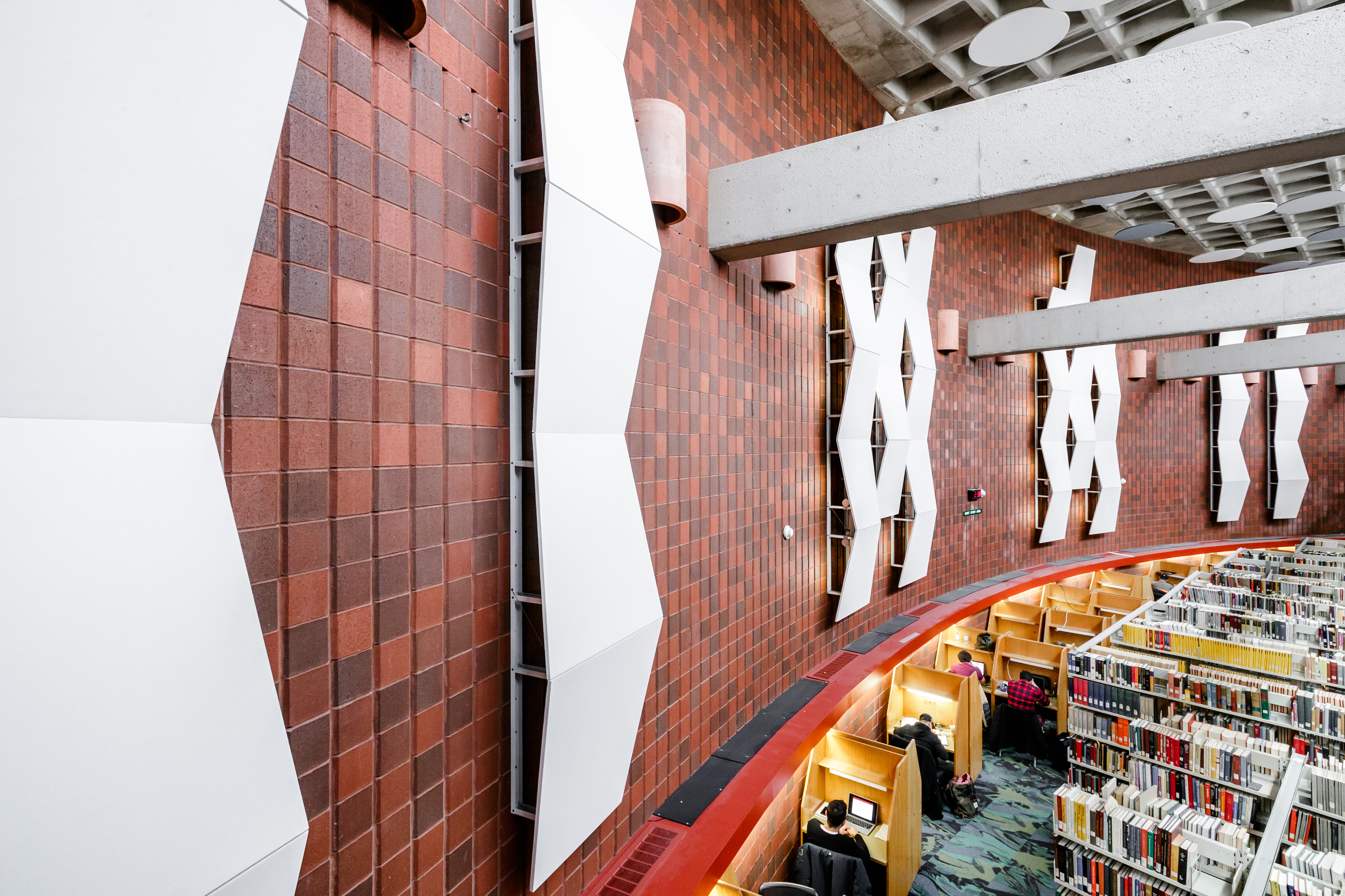 A row of study carrels lines the perimeter of the main floor in a library featuring red brick walls and large white panels.