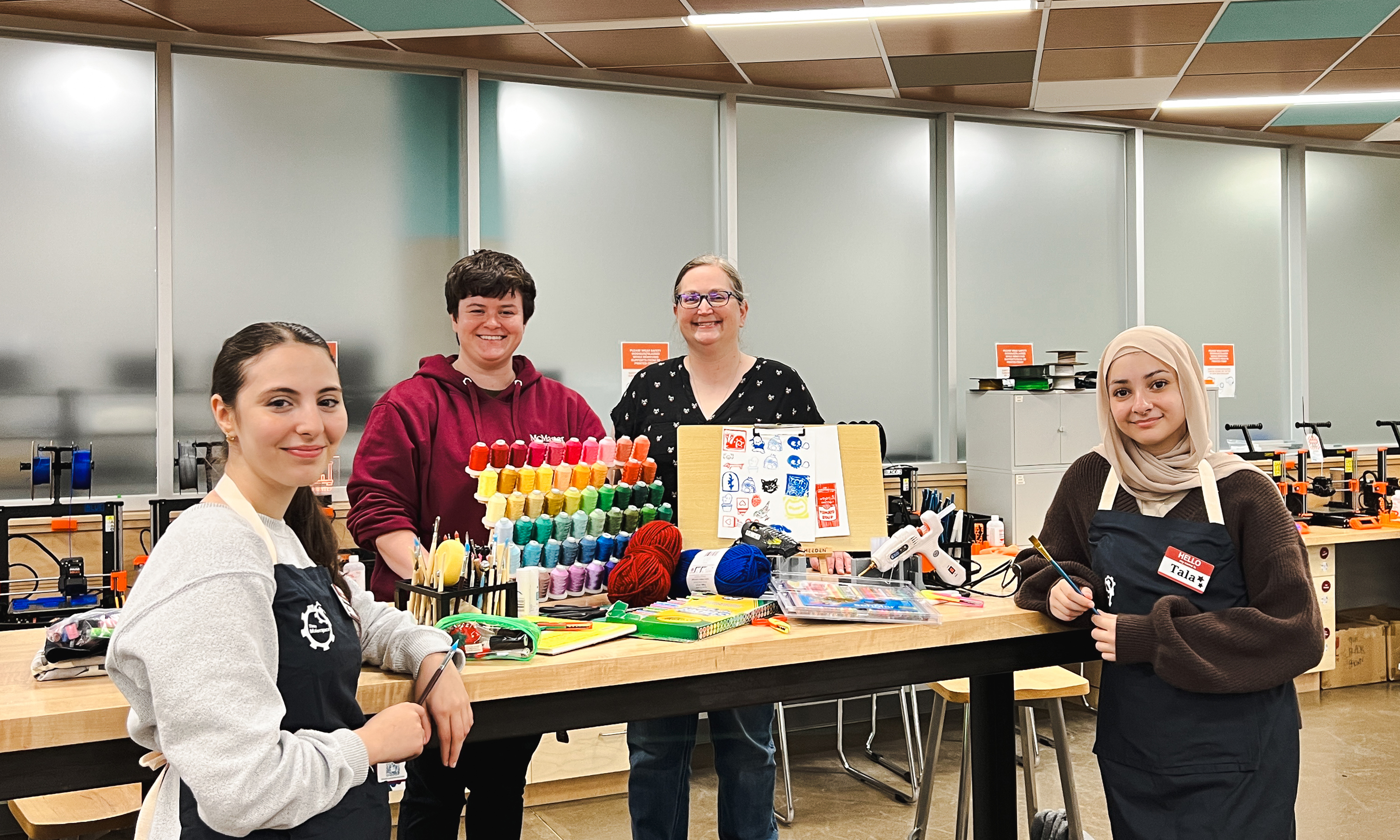Four libraries employees stand smiling around a long table featuring an assortment of crafting materials, including yarn, stamps, thread, markers, paintbrushes, and hot glue guns, inside Thode Makerspace.