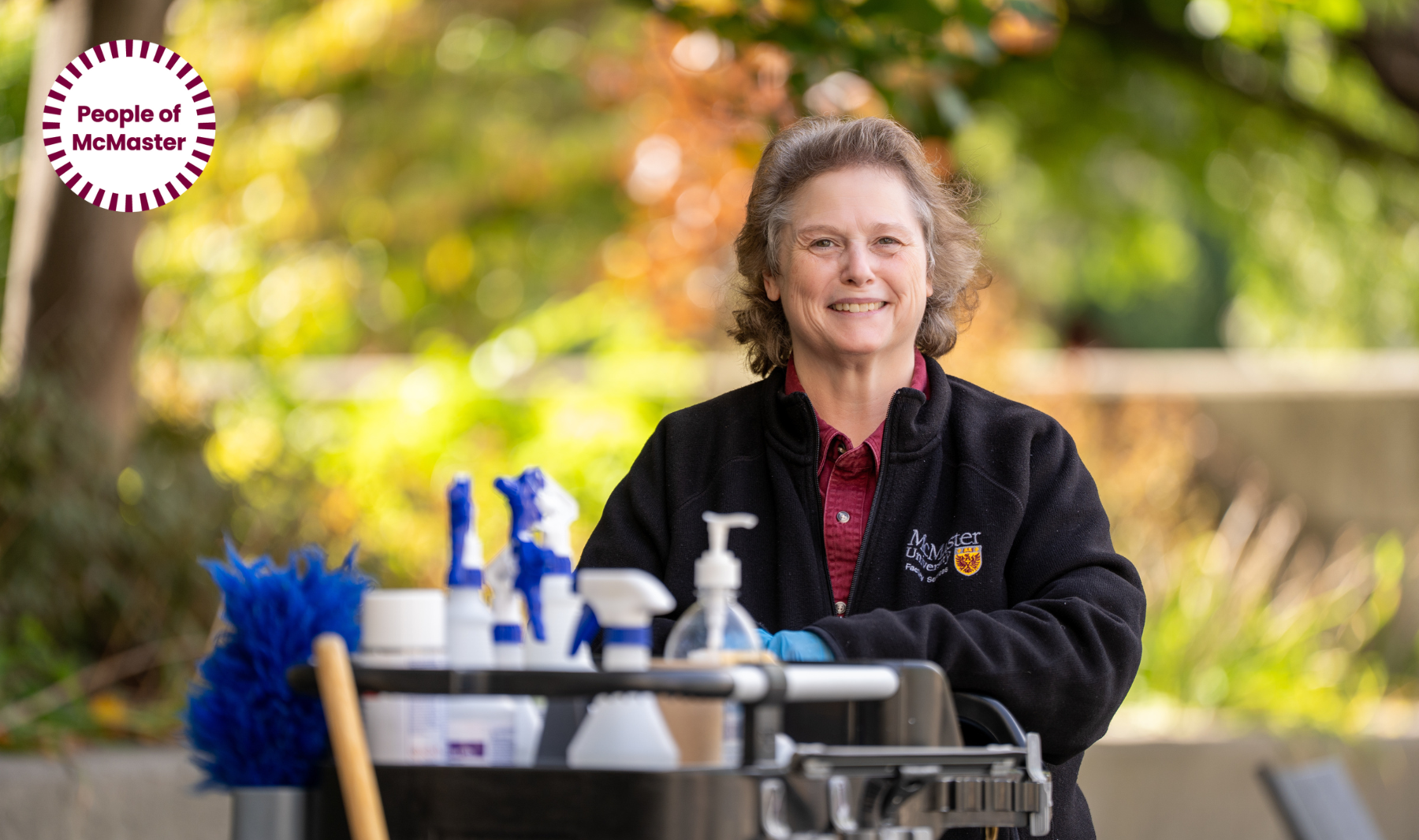 Tracie Large with short brown hair wearing a maroon shirt under a black McMaster jacket stands smiling with a cart of supplies outdoors.