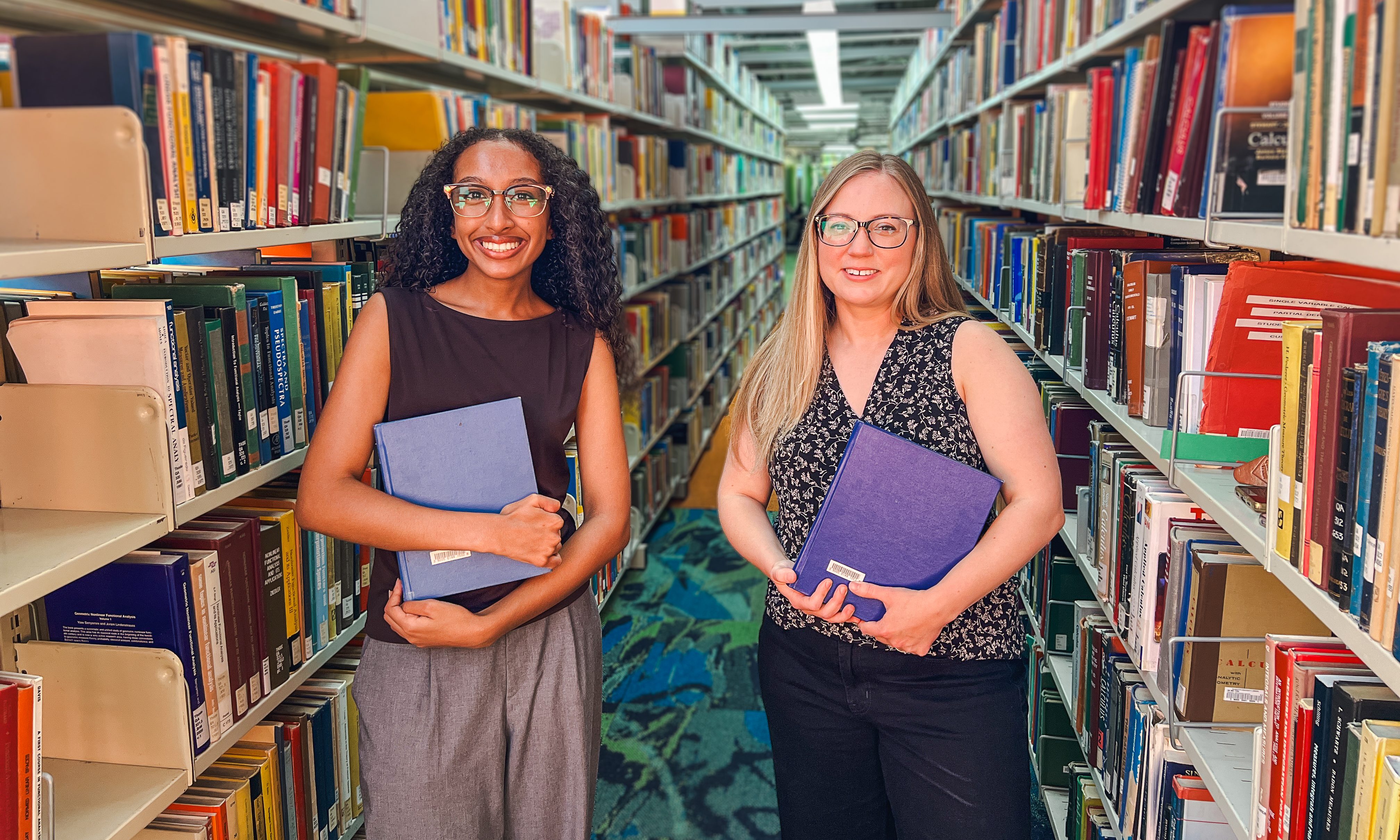 Rebecca with long black hair wearing glasses, black blouse and grey pants stands smiling beside Katie with long blonde hair wearing a black and white floral blouse and black pants also stands smiling, both holding textbooks between rows of bookshelves in a library.