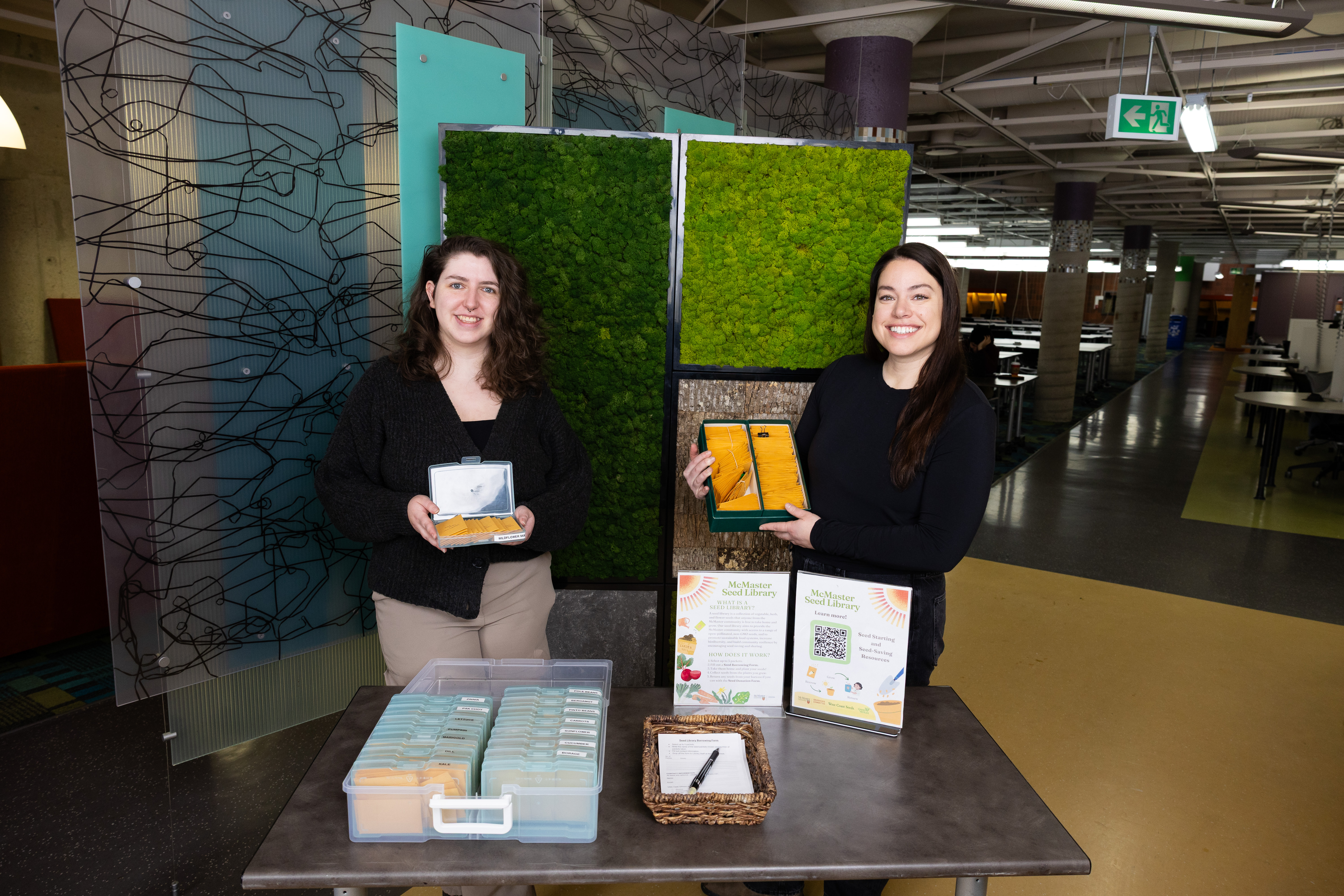 Paige with long brown hair and Nicole with long dark brown hair stand smiling while holding seeds in front of a moss wall inside the library.