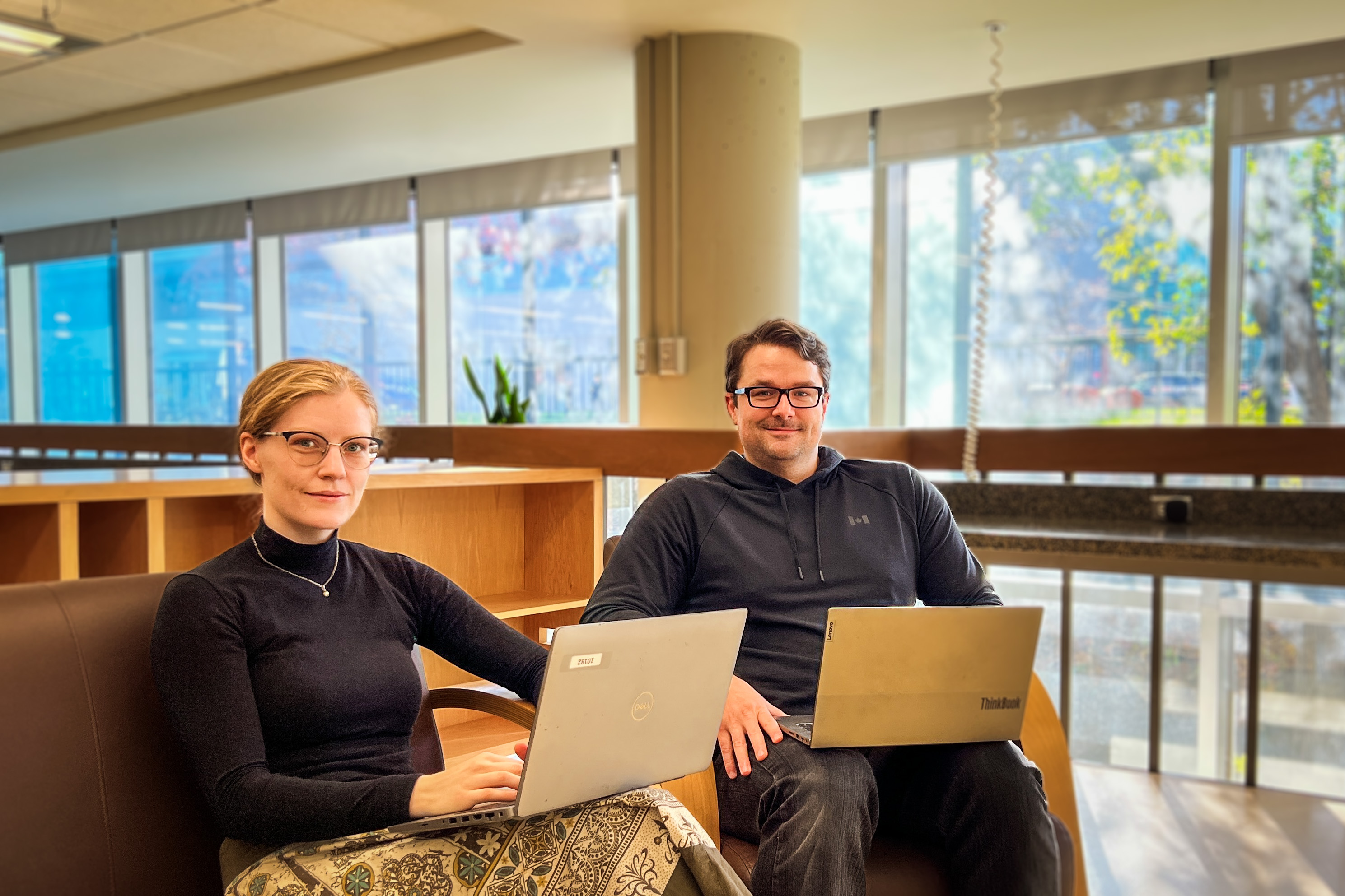 Two libraries employees sit smiling while working at laptops in a library space lined with windows.