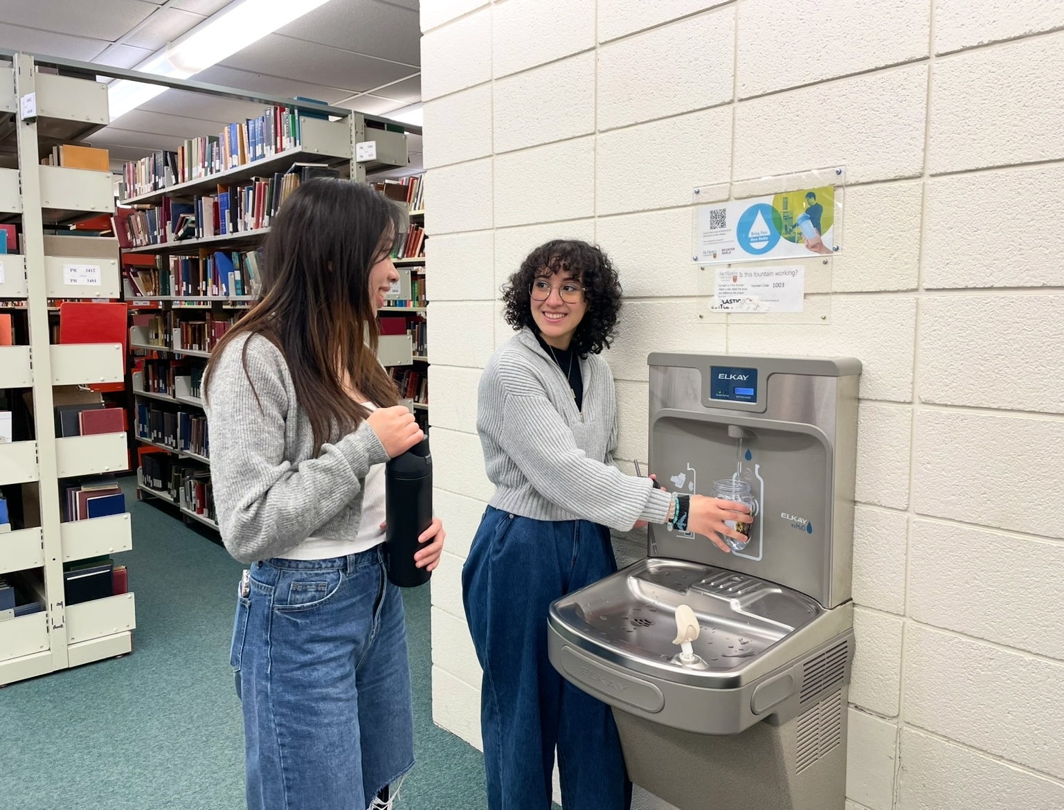 Rebecca with long brown hair wearing a grey sweater and jeans stands holding a reusable water bottle next to Victoria with short black hair wearing glasses, grey sweater and blue jeans, refilling a water bottle from a water fountain on a white brick wall with rows of bookshelves in the background. 