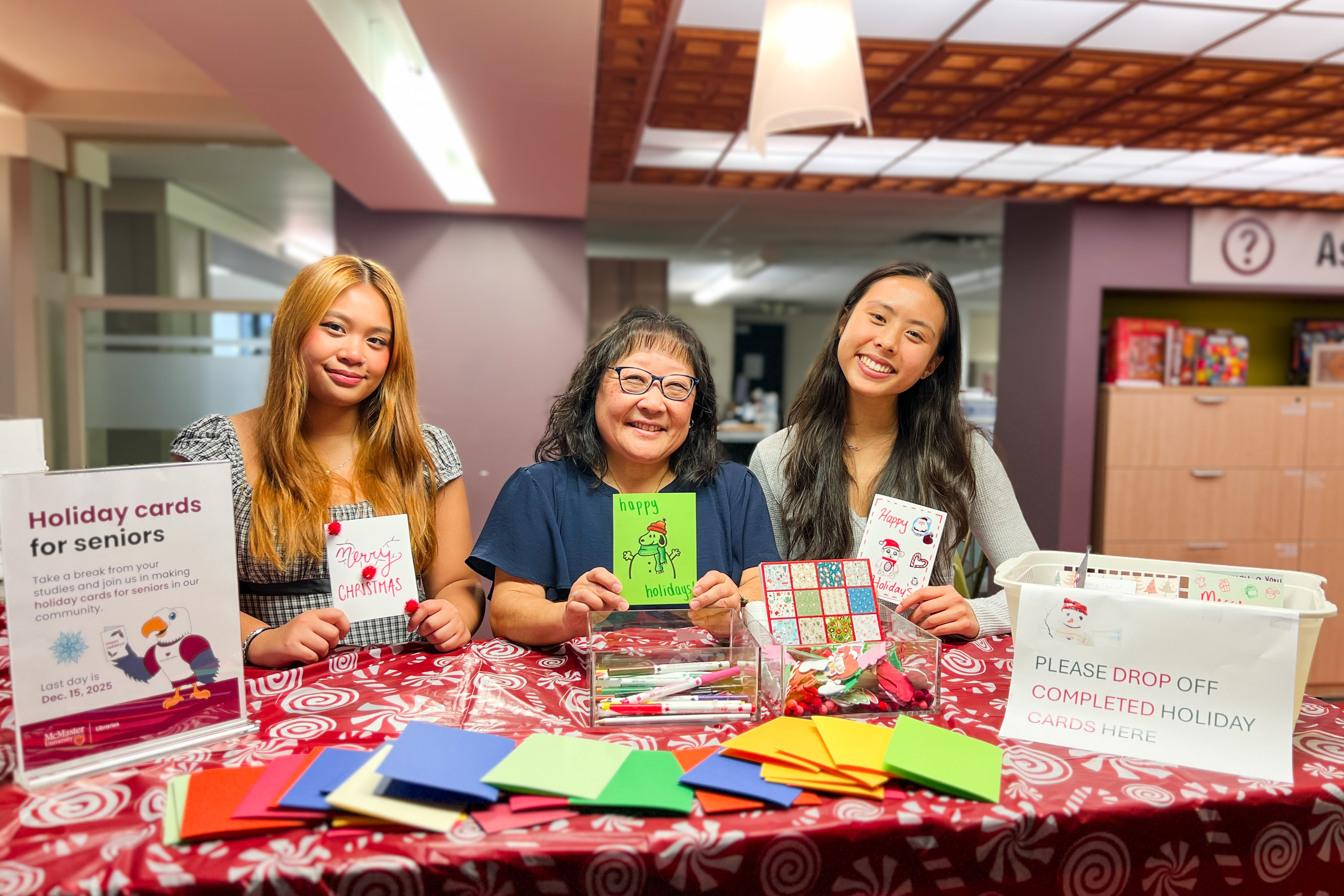 Two student assistants and one libraries manager stand together at a services desk in front of a display of paper, stickers and markers promoting holiday cards for seniors.