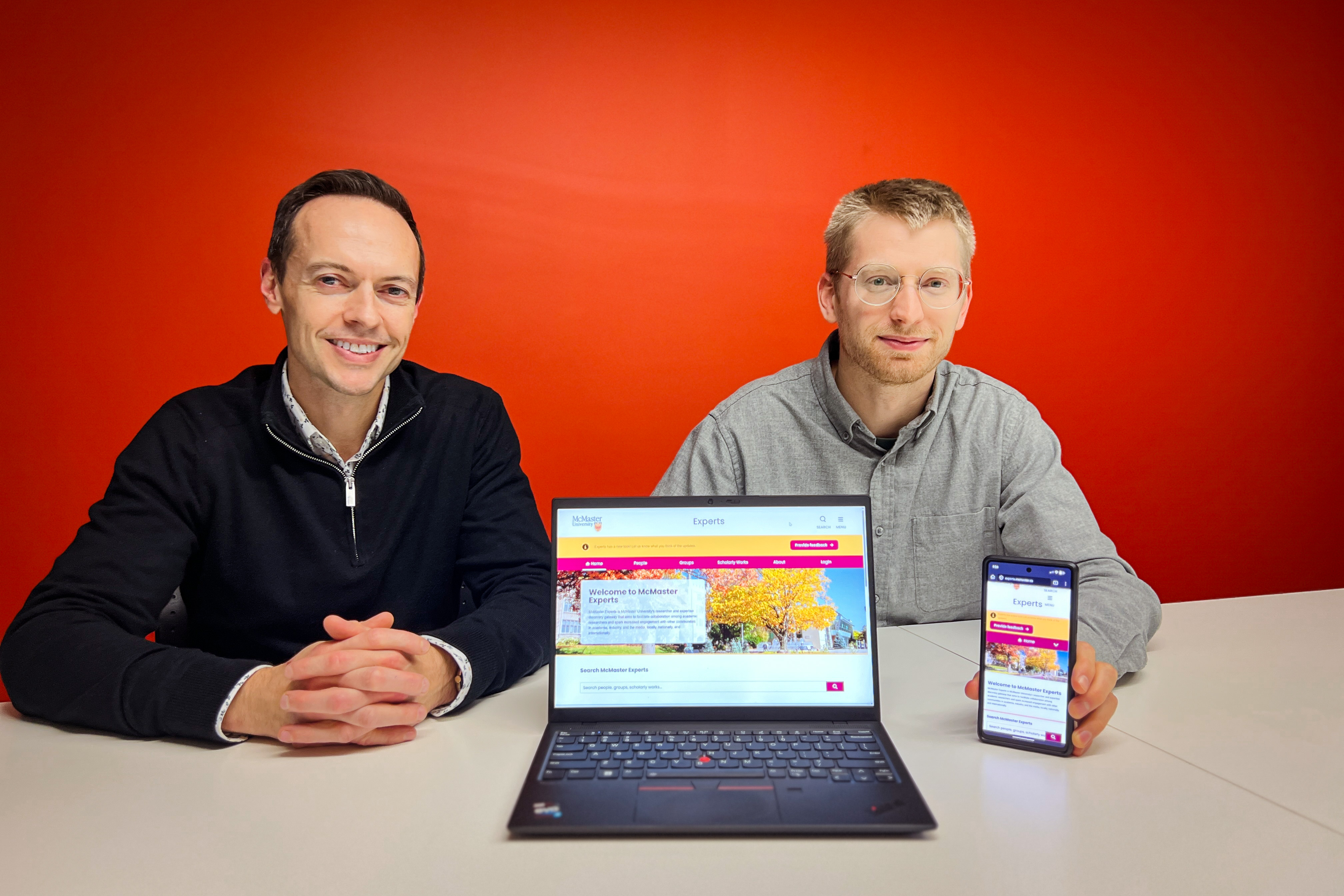 Jay with short black hair wearing a black sweater and Jacob with short blonde hair wearing glasses and a grey dress shirt holding a laptop and iPhone displaying the McMaster Experts sit at a white table in front of a red wall. 