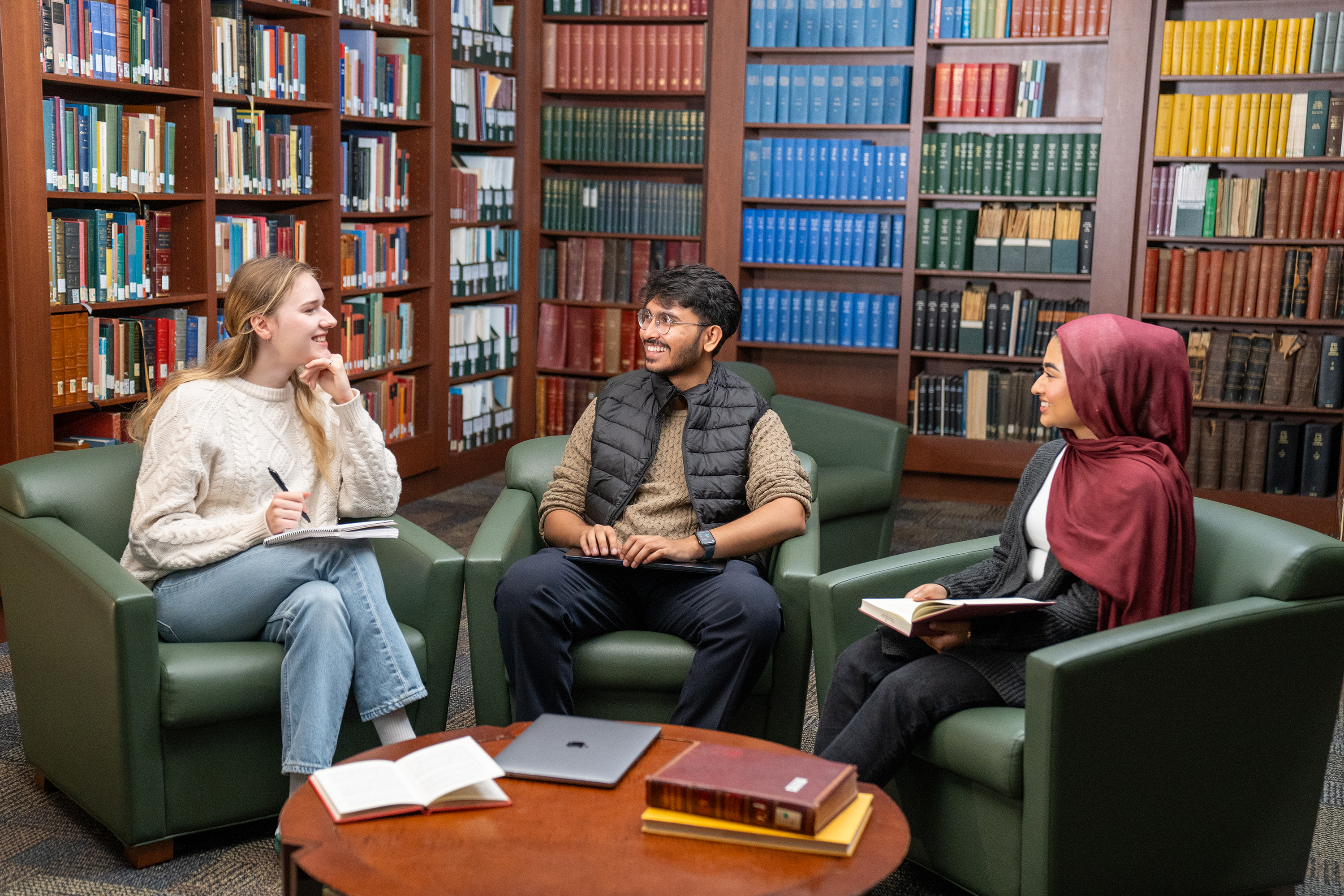 Three students sit with notebooks and pens in green armchairs in a wood paneled library room surrounded by stacks of books.