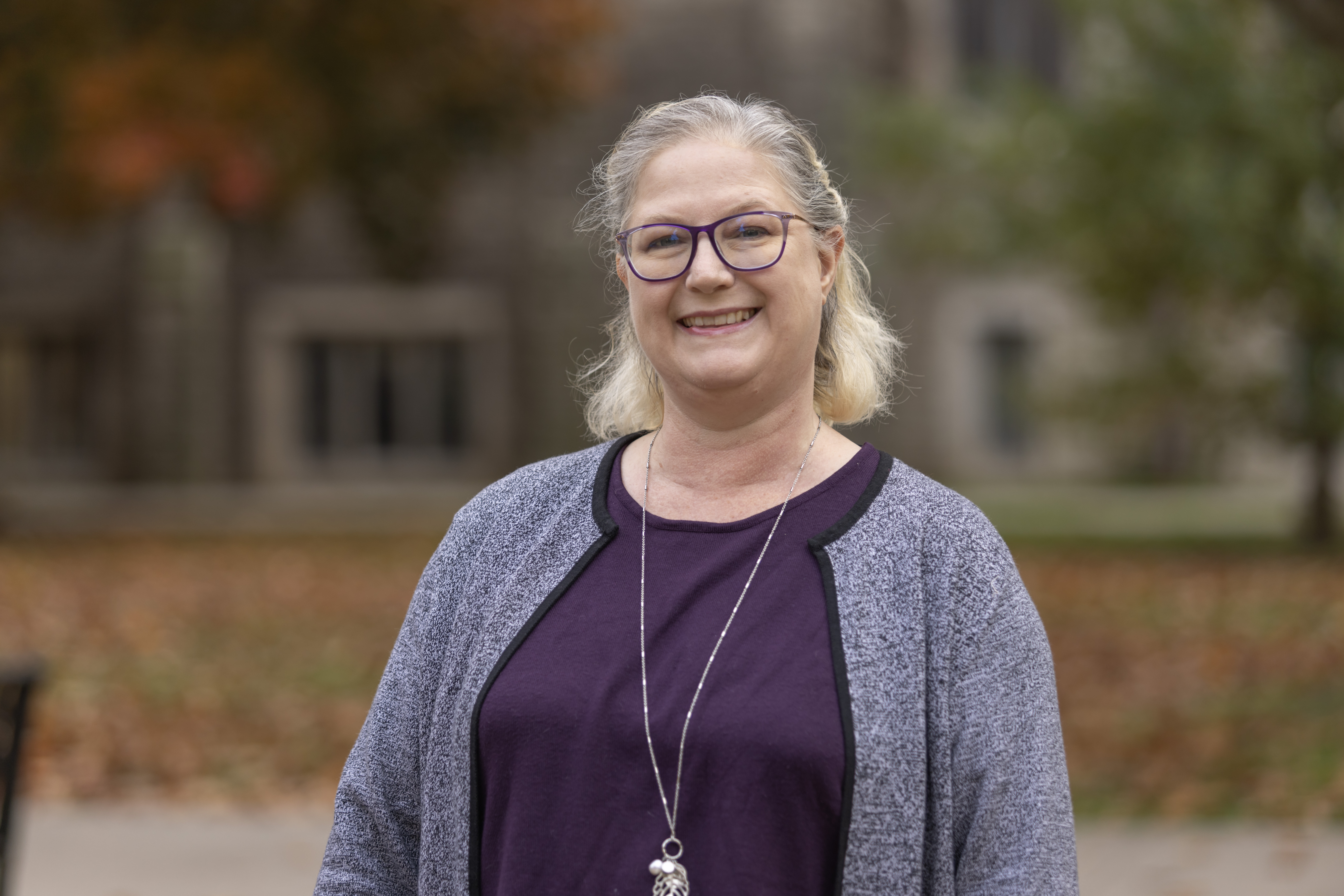 Cathy with shoulder-length blonde hair wearing glasses and a grey cardigan over a dark purple shirt stands smiling outside during a fall day on campus.