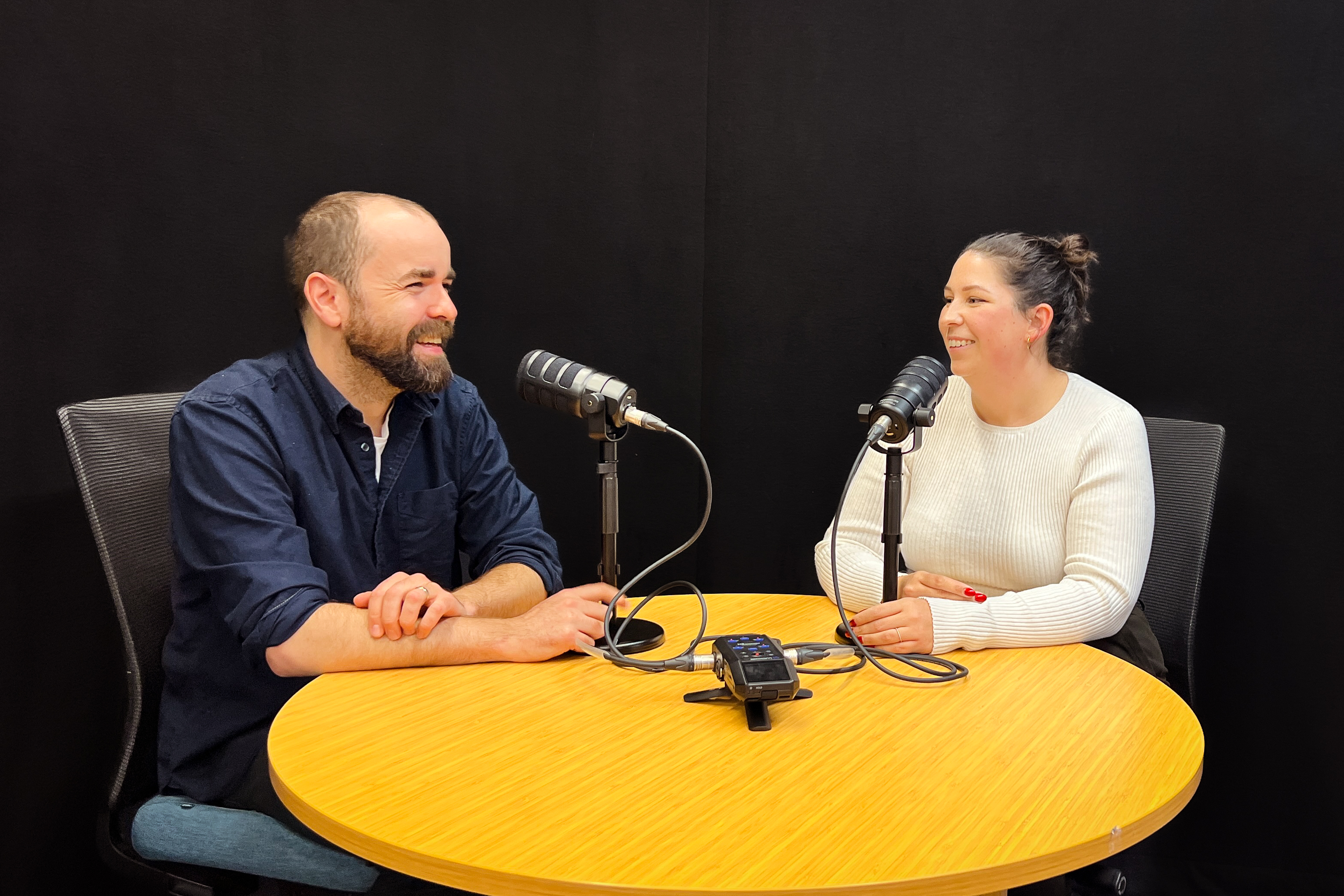 Jack with short brown hair and a beard wearing a blue collared shirt and Carlie with brown hair tied in a bun sit together at a wooden table with two microphones recording a podcast.