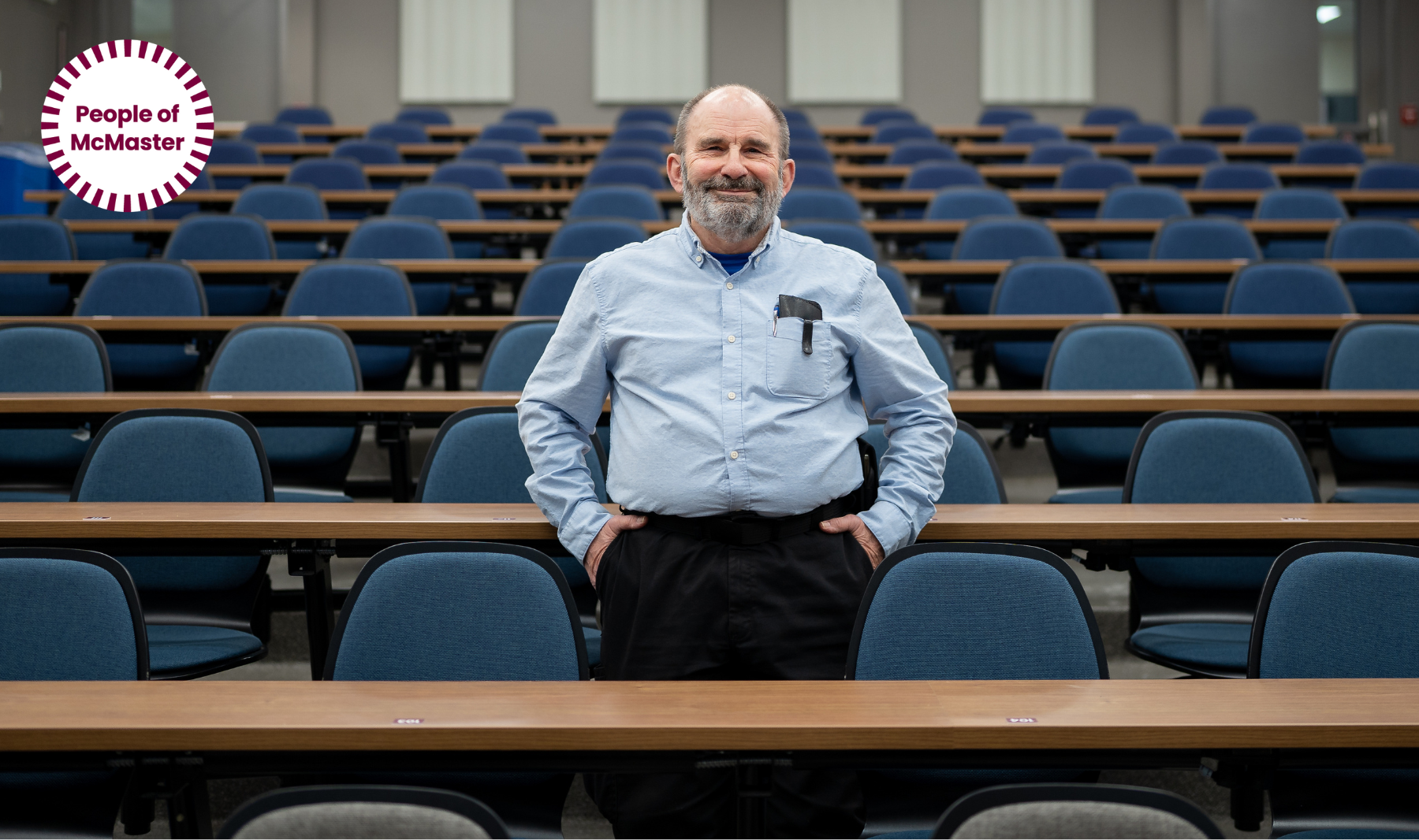 Kevin wearing a blue dress shirt and black pants stands smiling in an empty lecture hall at McMaster.