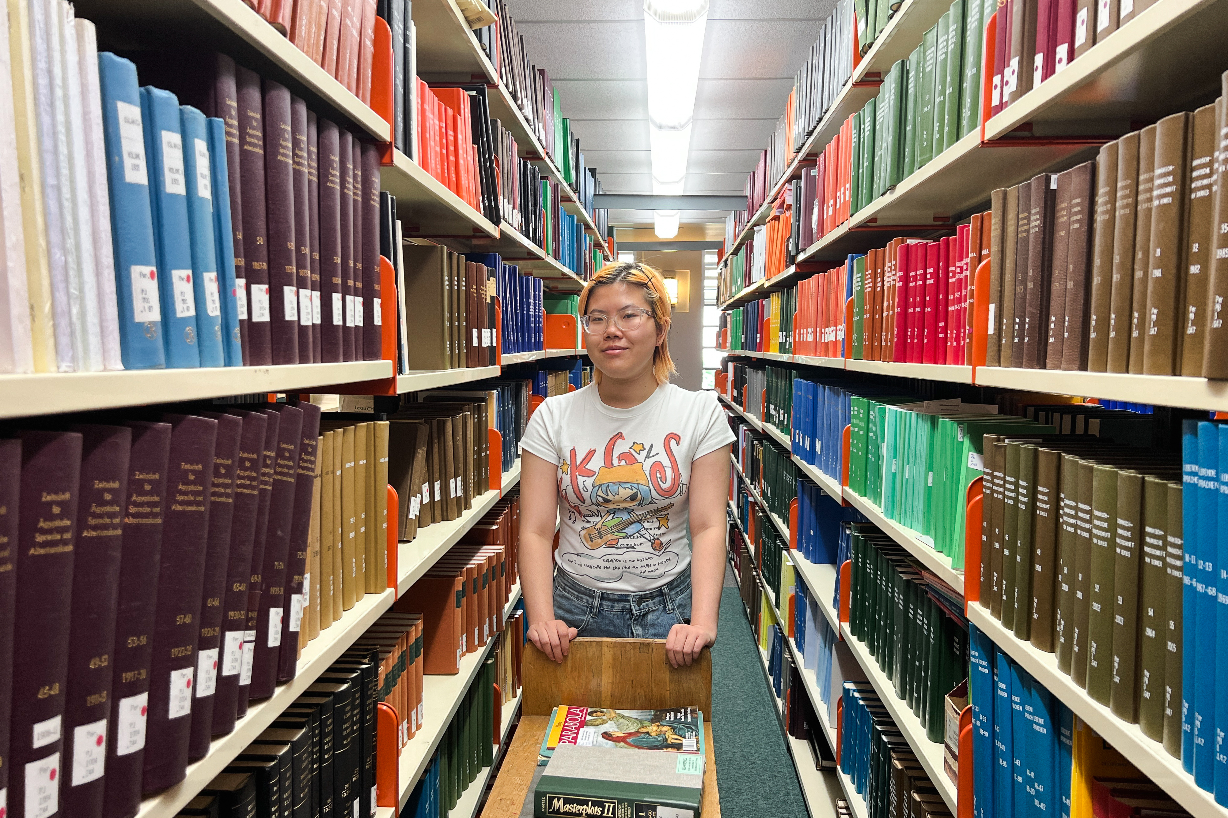Jane with short blonde hair and glasses wearing a white T-shirt stands behind a cart of library books. 