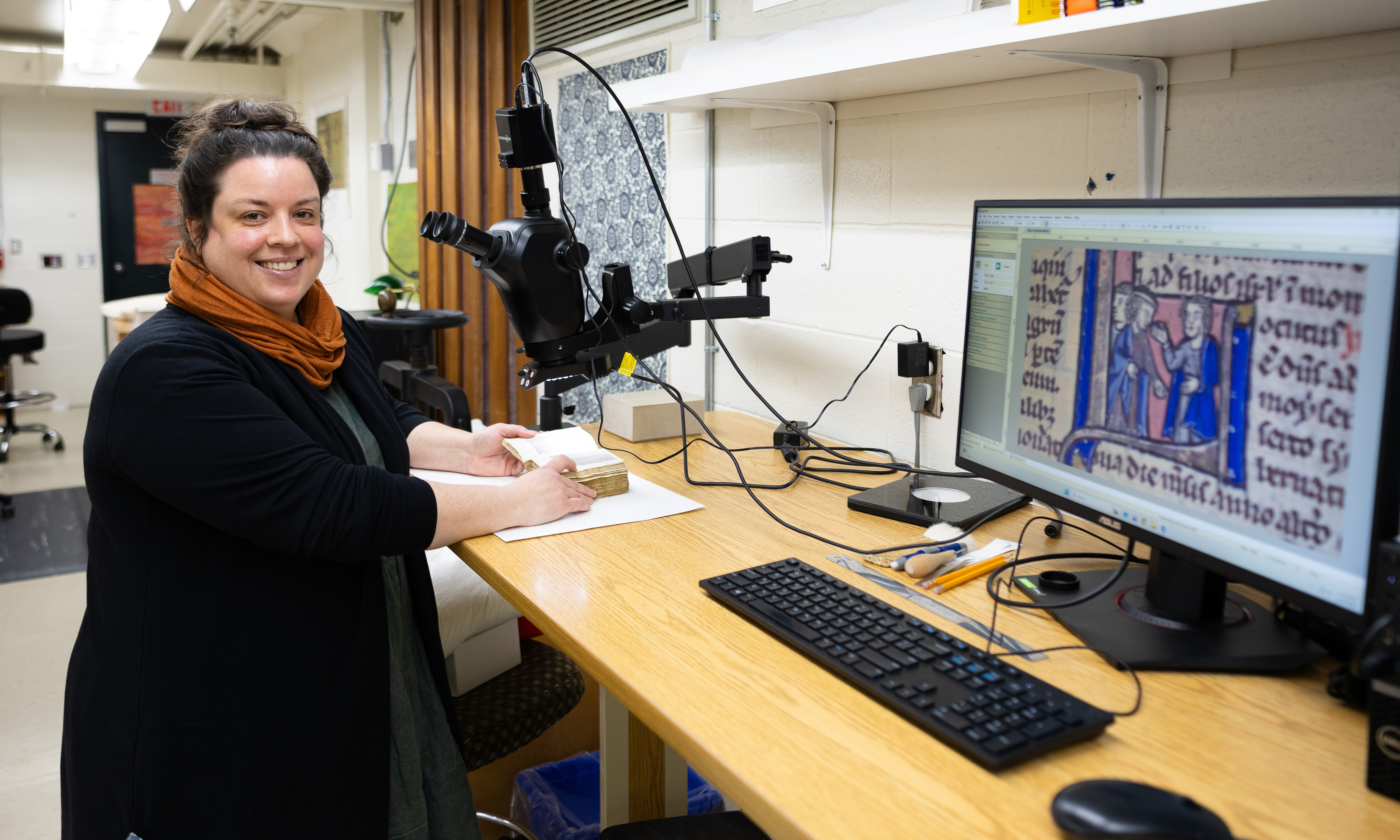 Bronwen with black hair in a bun wearing a black sweater and an orange scarf stands smiling at a microscope.