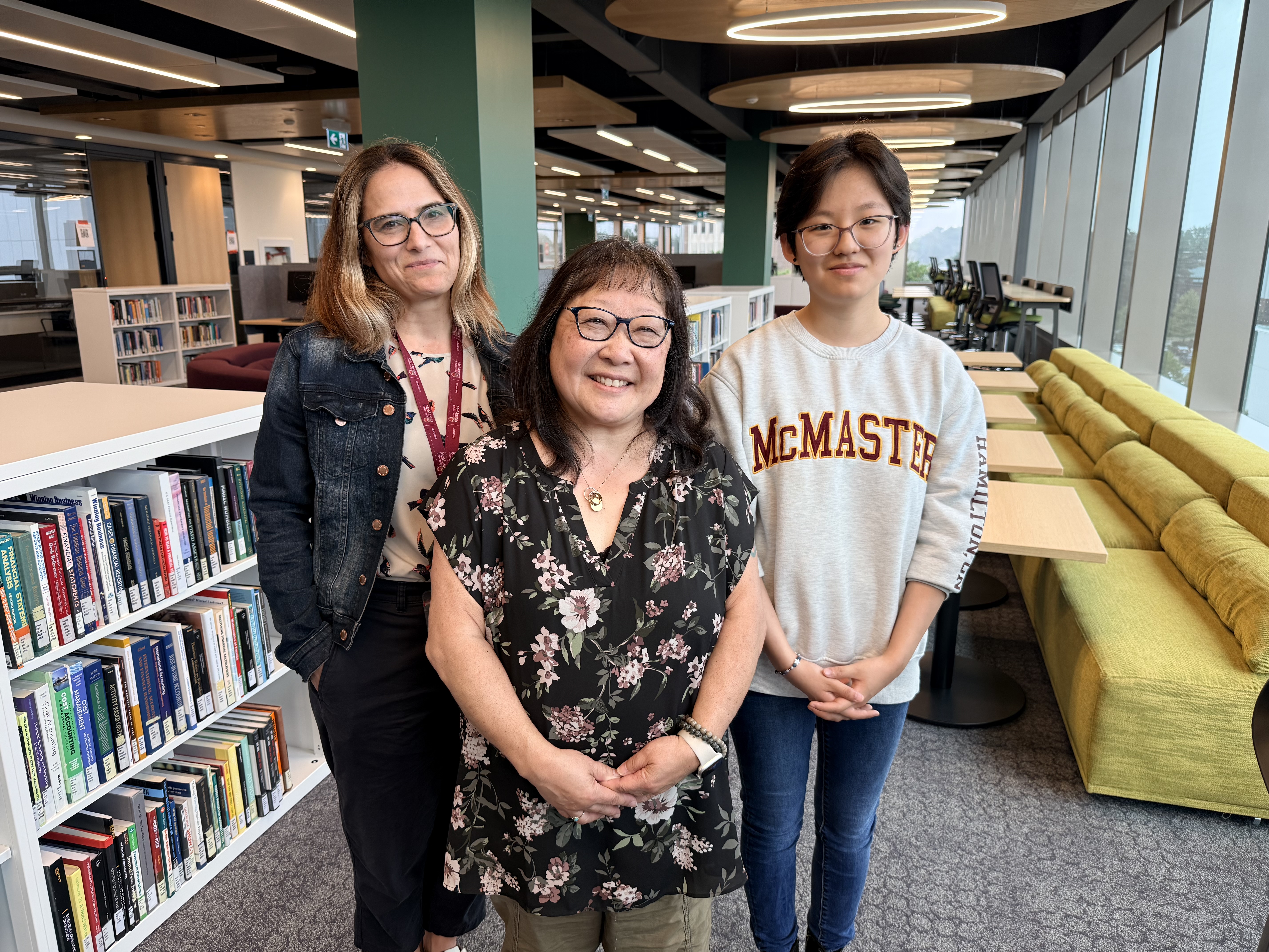 Lynn with brown hair wearing a jean jacket, Ann with glasses wearing a black floral blouse, and Erica with a grey McMaster sweater stand inside a new library featuring filled bookshelves and seating areas.