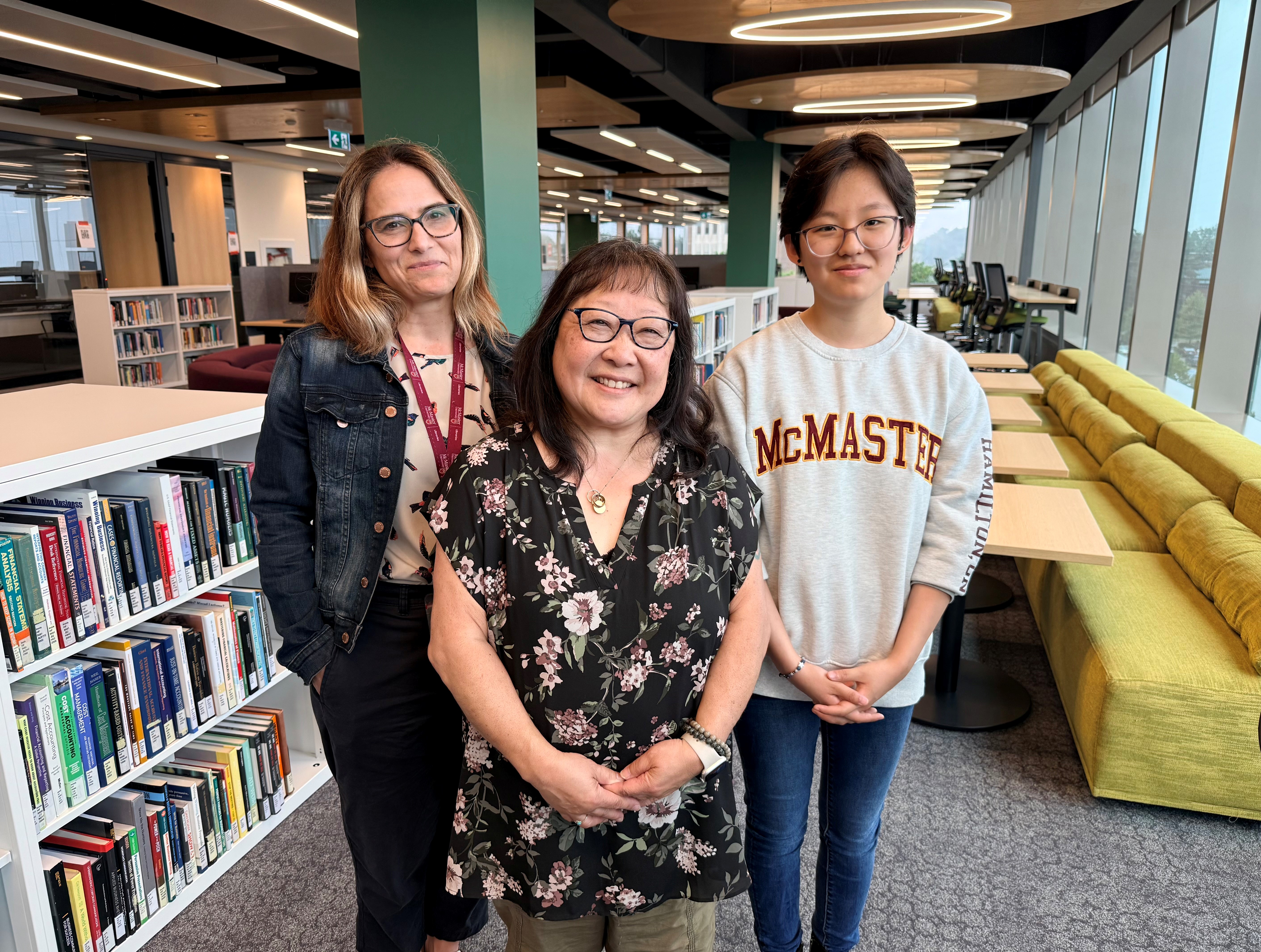 Lynn with brown hair wearing a jean jacket, Ann with glasses wearing a black floral blouse, and Erica with a grey McMaster sweater stand inside a new library featuring filled bookshelves and seating areas.