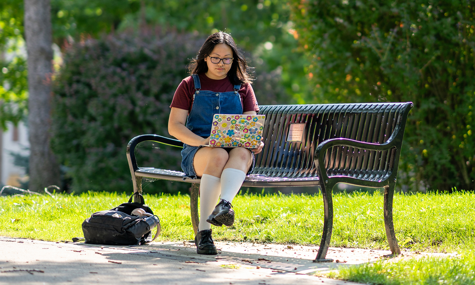 A student with black hair wearing glasses, a maroon shirt and denim overalls sits outdoor on a black metal bench on a sunny summer day.