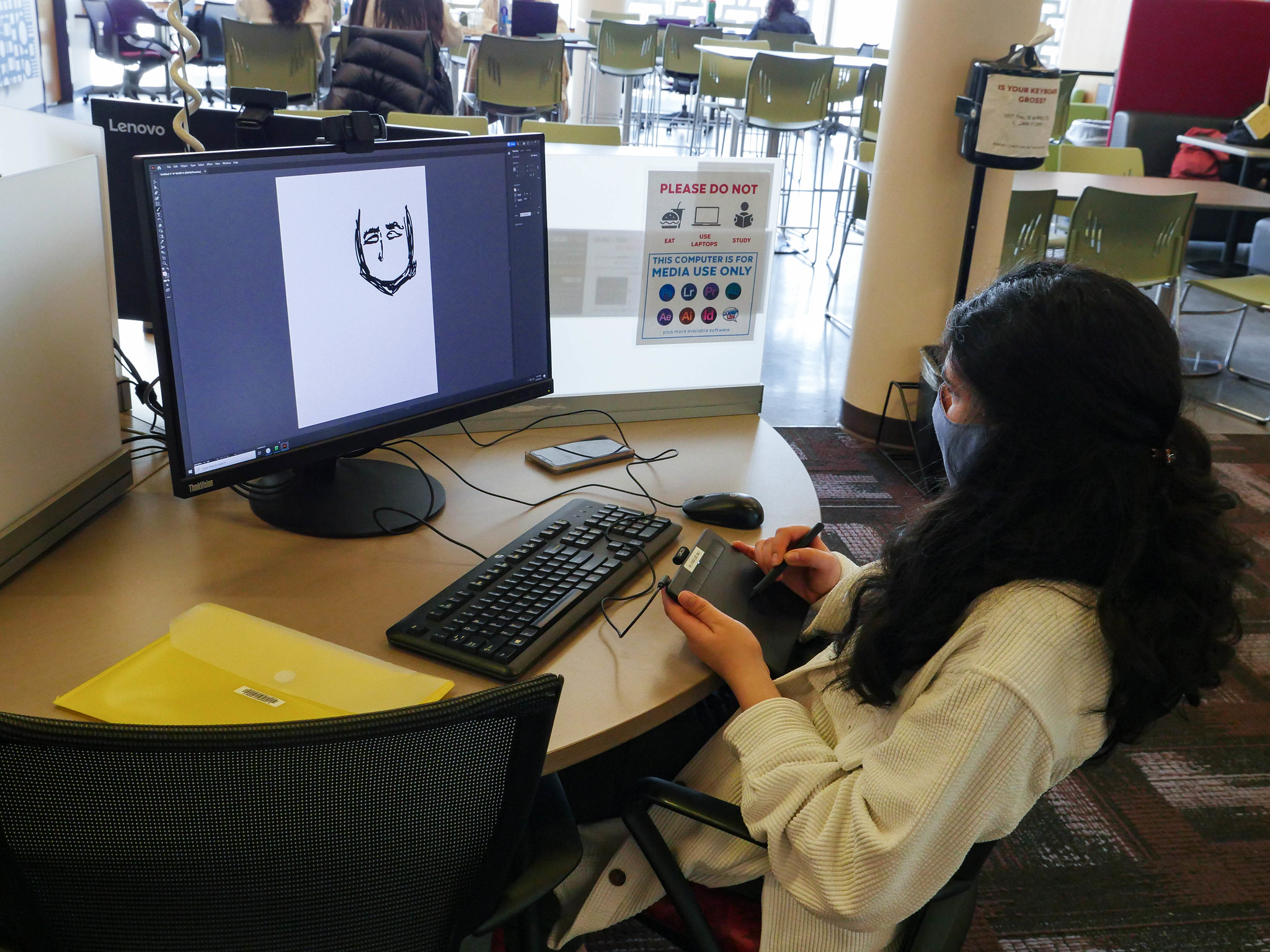 A student wearing a mask uses a digital art tablet to sketch a face on the computer in front of her.
