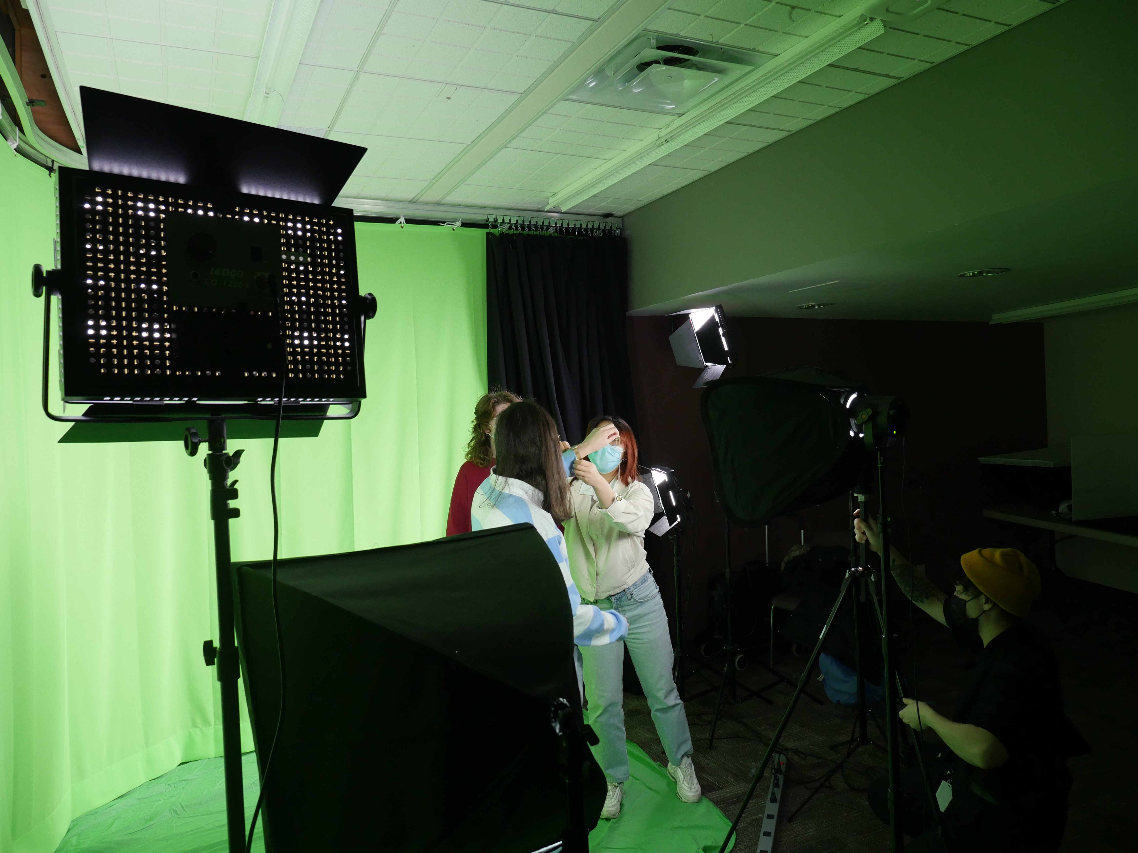Students stand in front of a green screen while another student adjusts the camera lighting.