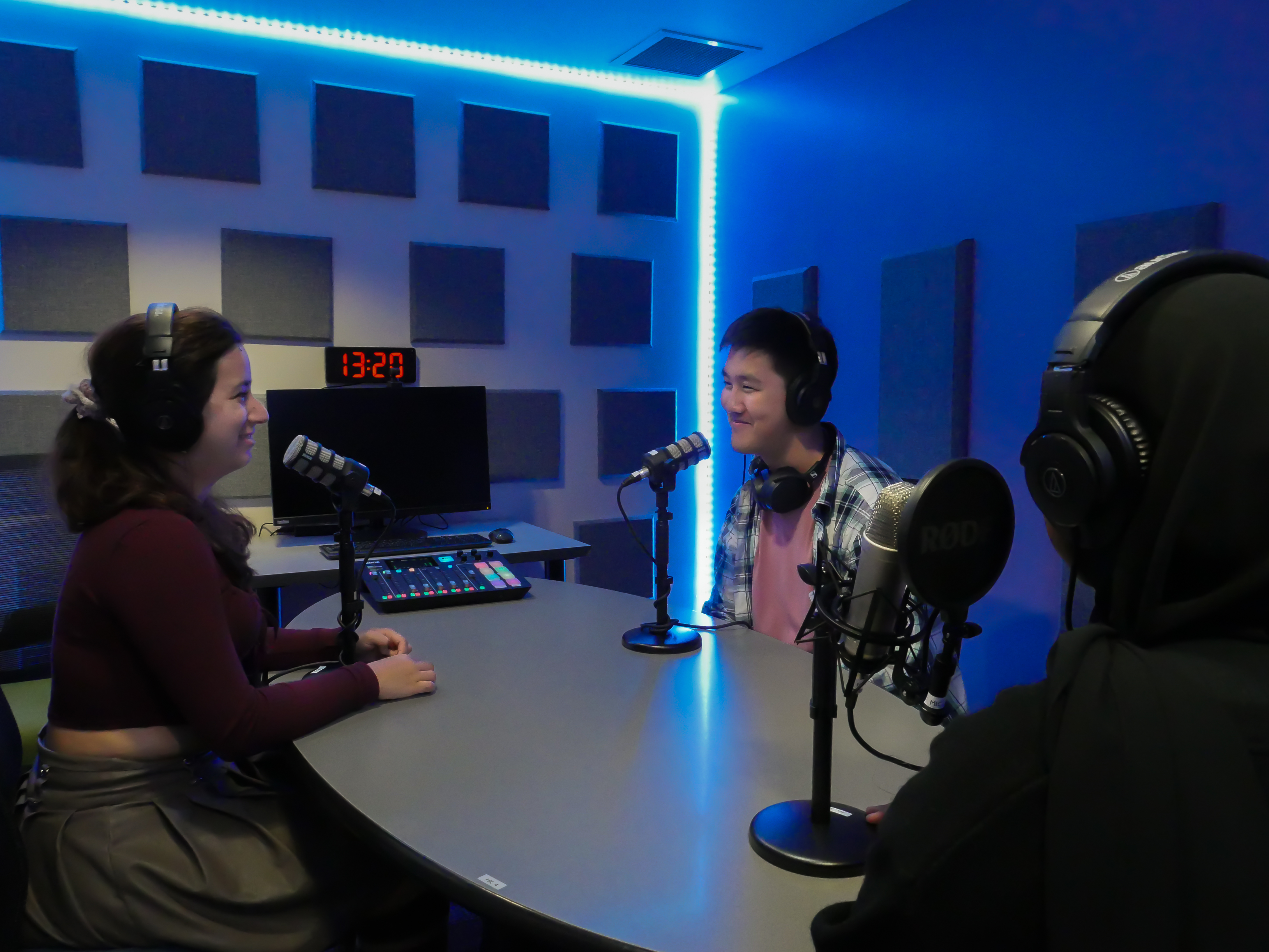 Students sit around a table in the podcast studio with LED lighting & soundproofing panels on the walls. Each are wearing headphones and are speaking into podcast microphones.