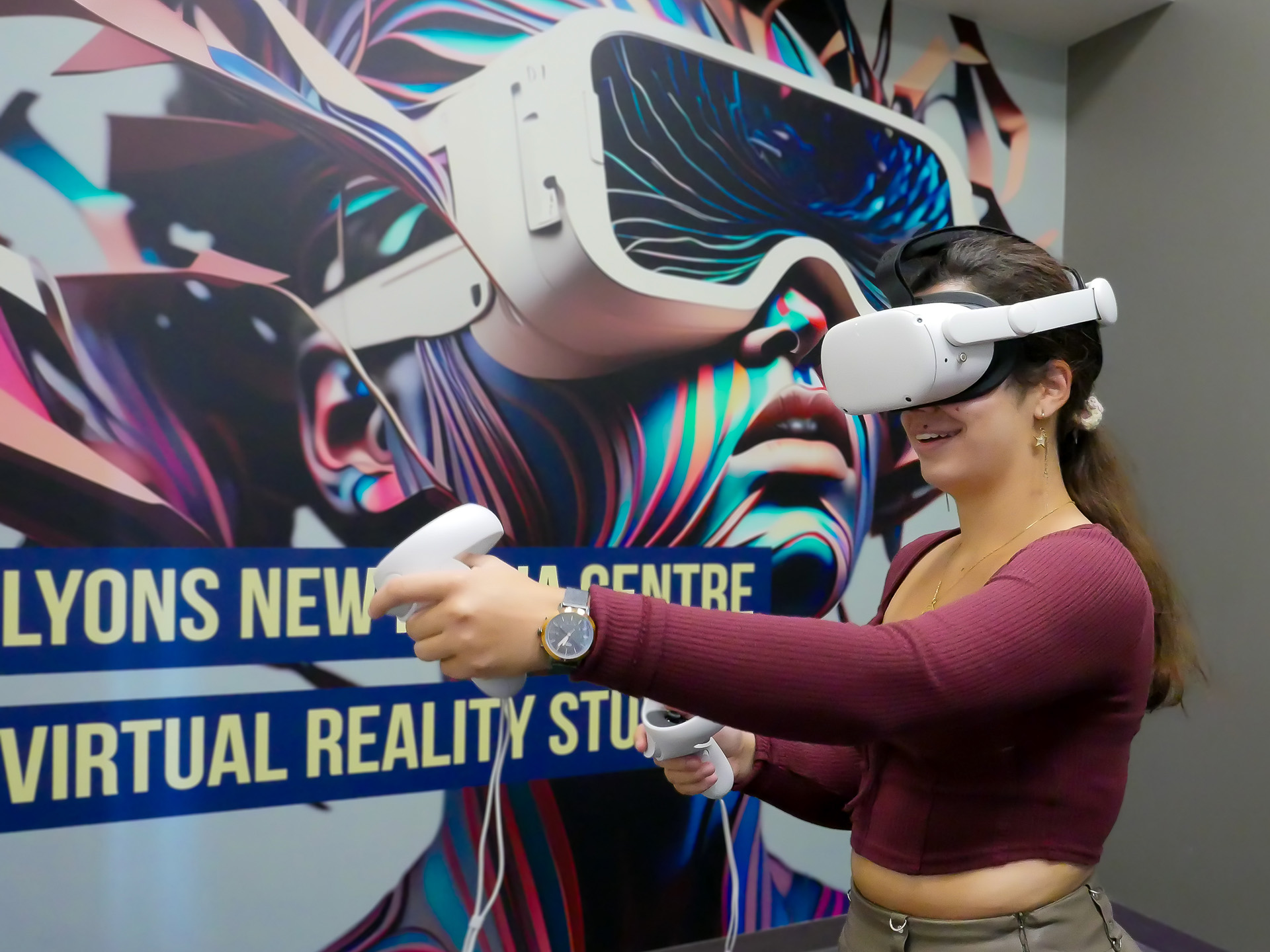 A young woman wearing a VR headset and holding the controllers tries out the virtual reality room in Lyons. A colourful, futuristic image of a woman wearing VR goggles can be seen on the wall behind.